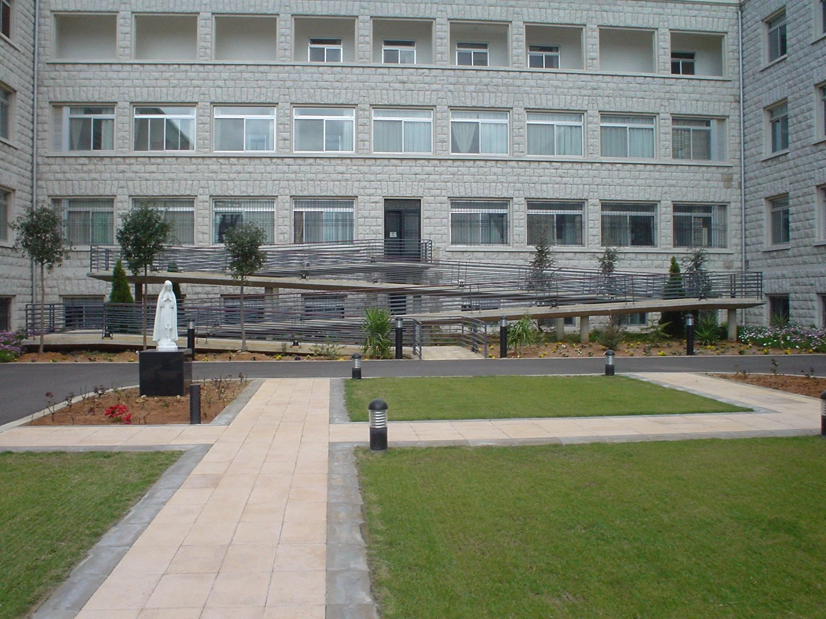 A courtyard with a walking path, green grass, and small flower beds against a gray stone building. There is a statue and a small ramp with black railings for accessibility.