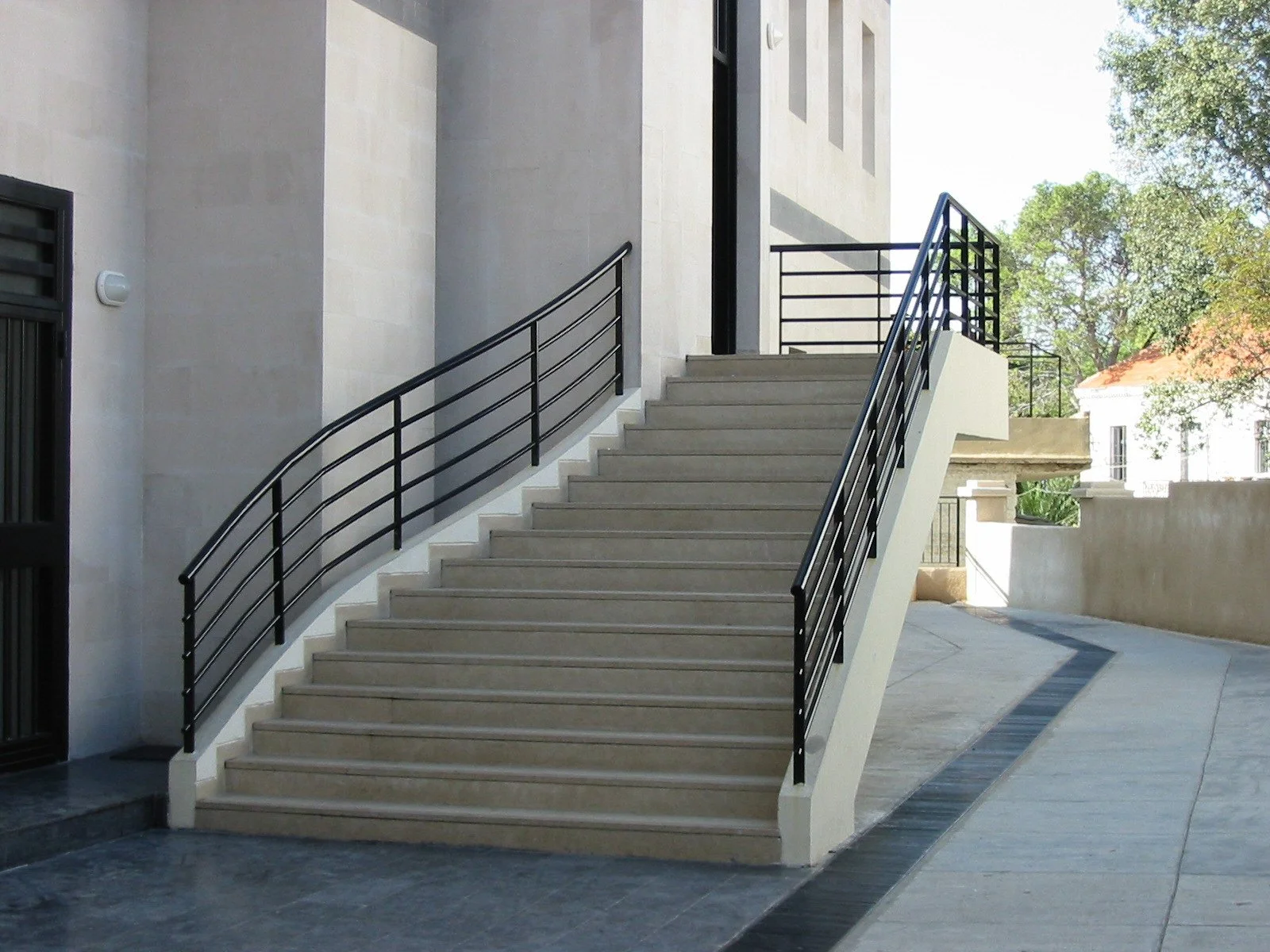 Exterior staircase with beige steps and black metal railing, leading up to a building entrance, with trees and another building in the background.