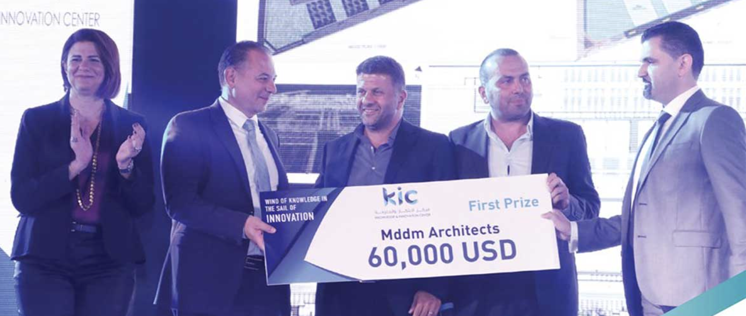 A group of five people on stage holding a large check for $60,000 USD awarded to Mddm Architects, with the text 'First Prize' and 'Wind of Knowledge in the Sail of Innovation'. The background has bright screens and a cityscape view.