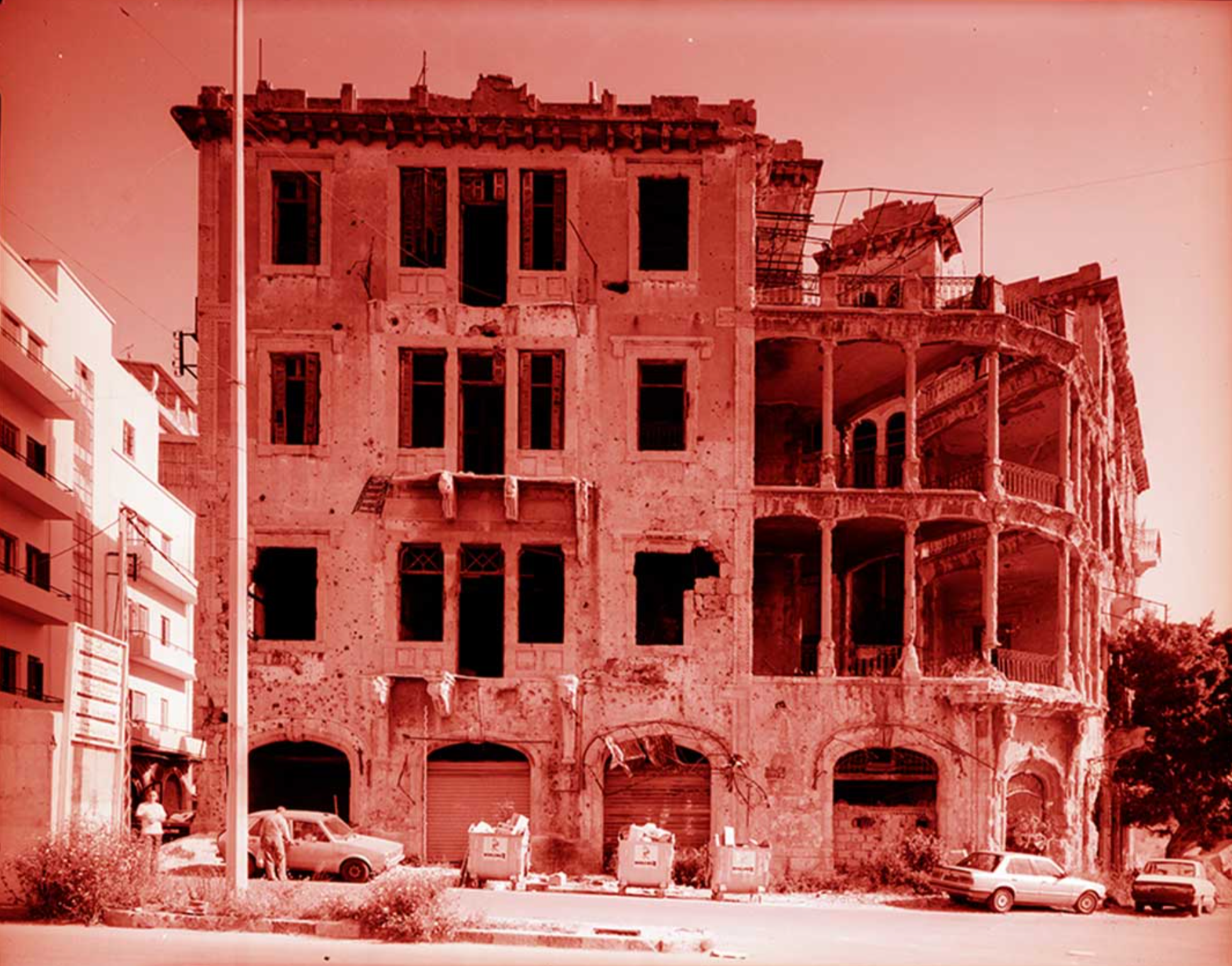 An old, worn-out building with broken windows and damaged walls, located on a street with parked cars and a few pedestrians.