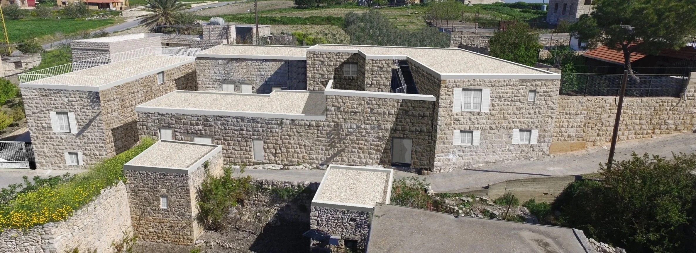 An architectural rendering of a proposed stone residential complex in Derdghaya Village, designed without roof tiles, featuring multiple interconnected buildings with stone walls, small windows, and flat roofs, surrounded by greenery and a dirt road.