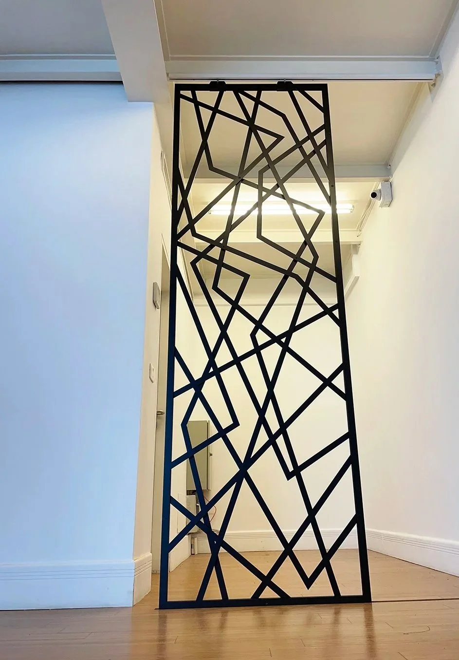 A tall black metal room divider with an abstract geometric pattern, placed in a corridor with wooden flooring and white walls.