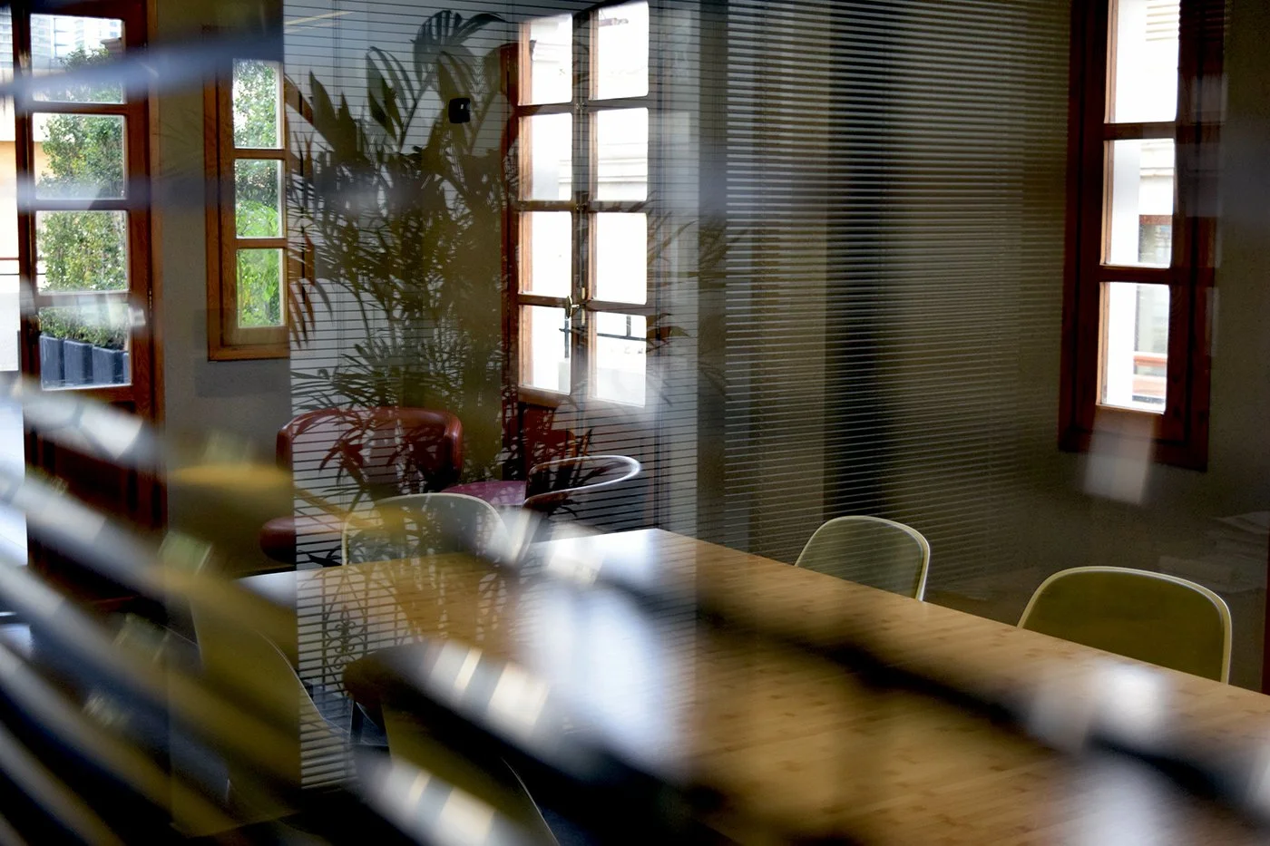 Interior of a room viewed through partially closed window blinds, featuring a wooden table, chairs, a plant, and multiple windows allowing natural light.