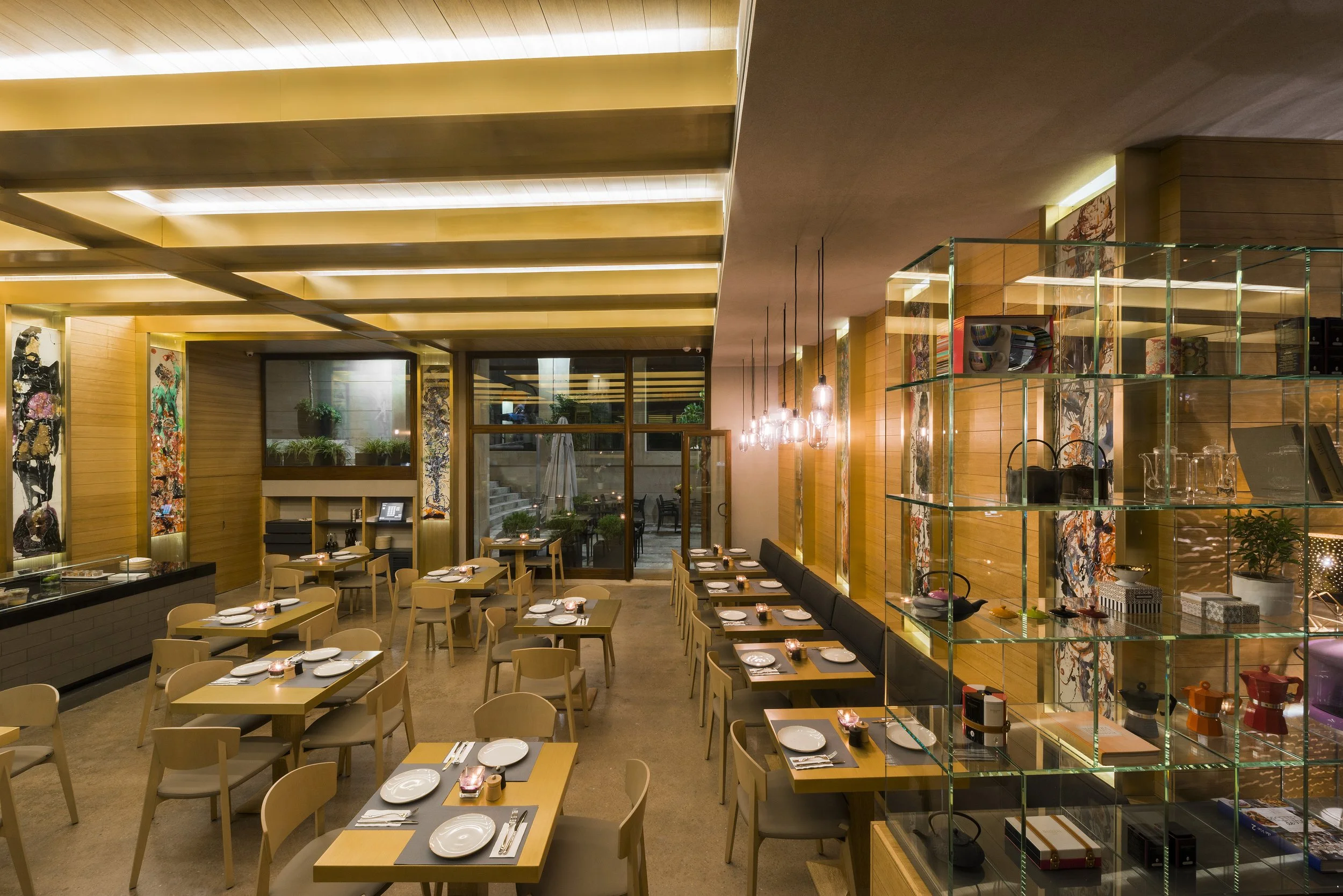 Interior of a modern restaurant with wooden walls, tables set with plates and cutlery, warm lighting, and decorative artwork. Large glass display shelves hold various items, and large windows overlook an outdoor patio.