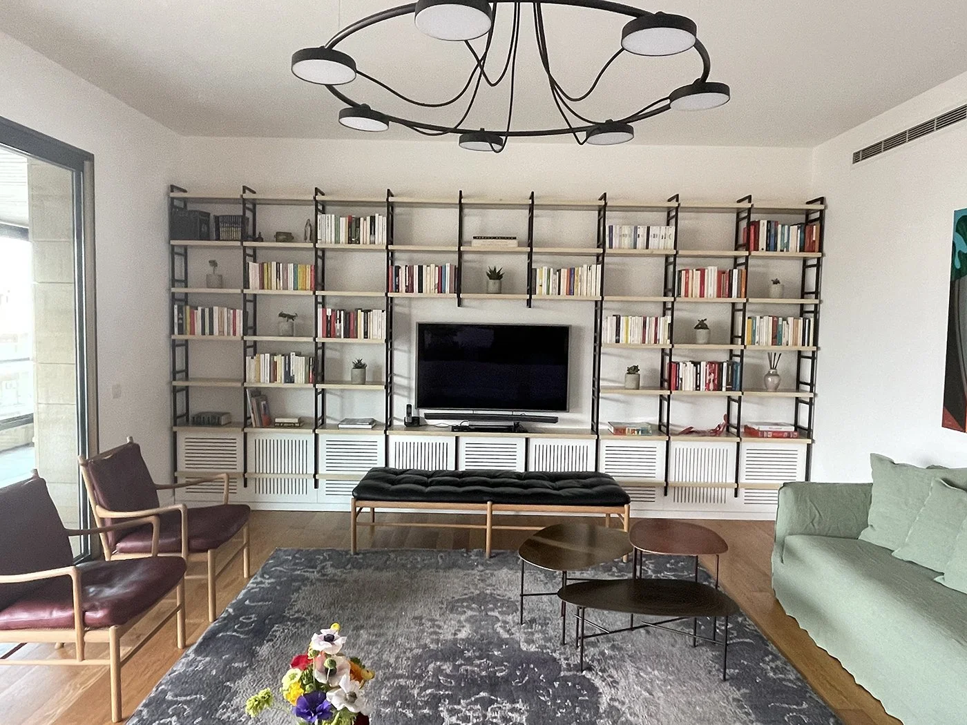 Living room with a large black-and-white bookshelf behind a flat-screen TV. There are two green pillows on a light green sofa and two wooden chairs with maroon cushions. A dark tufted bench and a set of three nested coffee tables are in front of the sofa. A gray rug with a floral pattern is on the wooden floor, and a colorful flower arrangement is on the rug. A sliding glass door leads outside on the left side, and there's a black modern chandelier overhead.