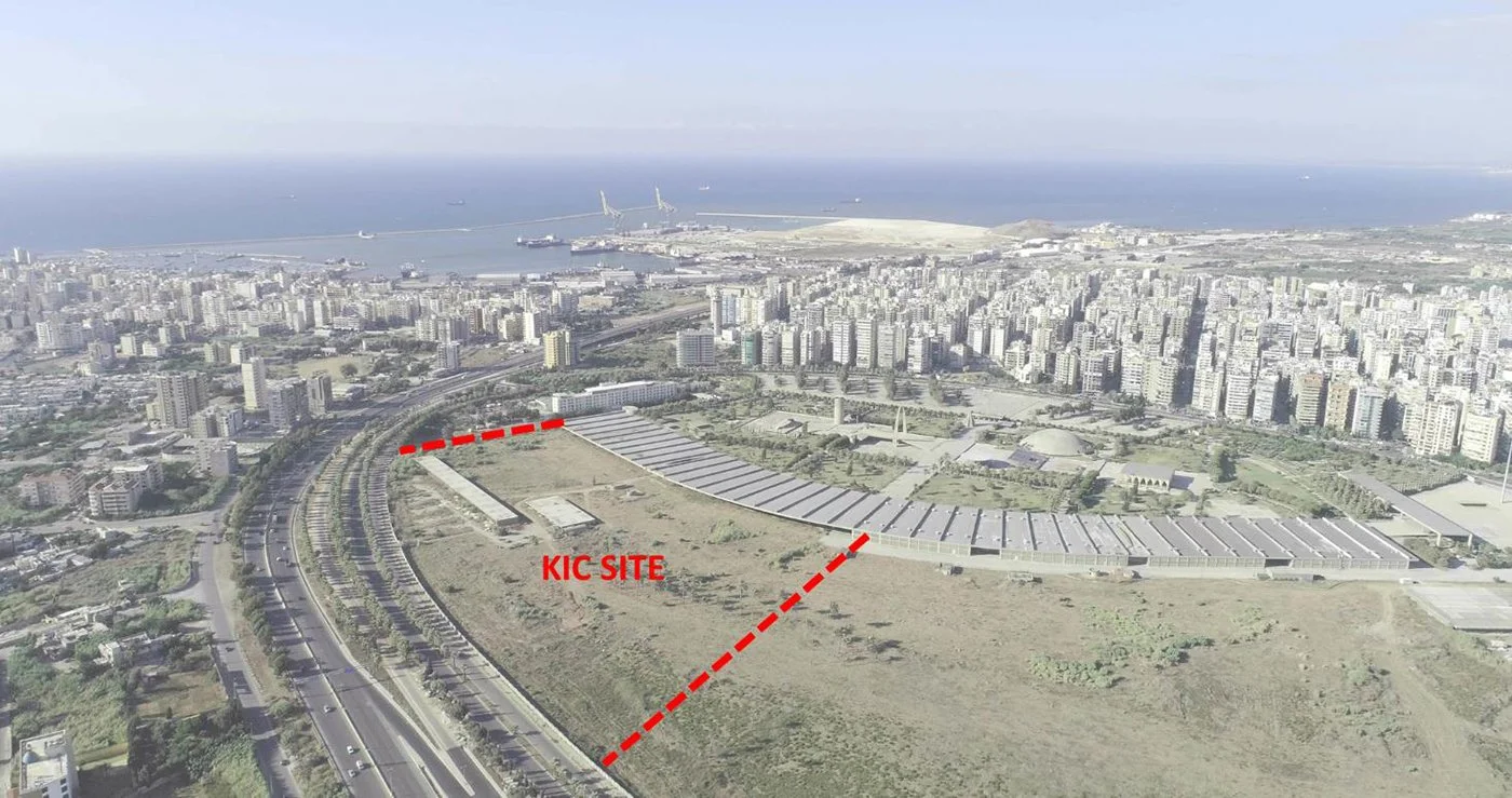 Aerial view of a city with numerous high-rise buildings, a large body of water in the background, and a construction site marked as 'KIC SITE' with a red dashed line. The site is near a highway with multiple lanes.