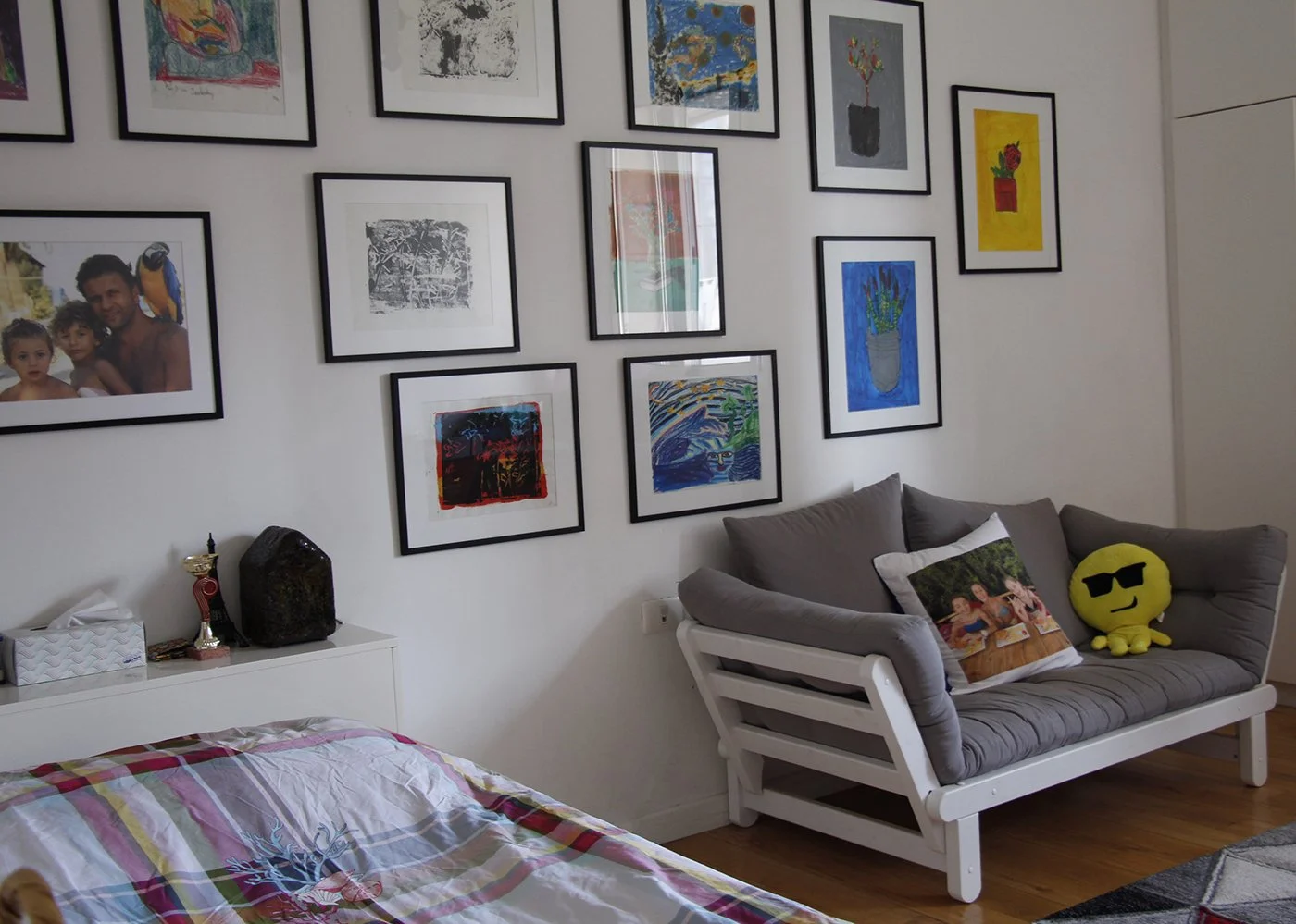 A living room corner with a grey couch decorated with cushions and plush toys, a gallery wall with framed artwork and photographs, a white side table with a trophy, tissue box, and a sculpture, and a bed with a colorful striped blanket in the foregro