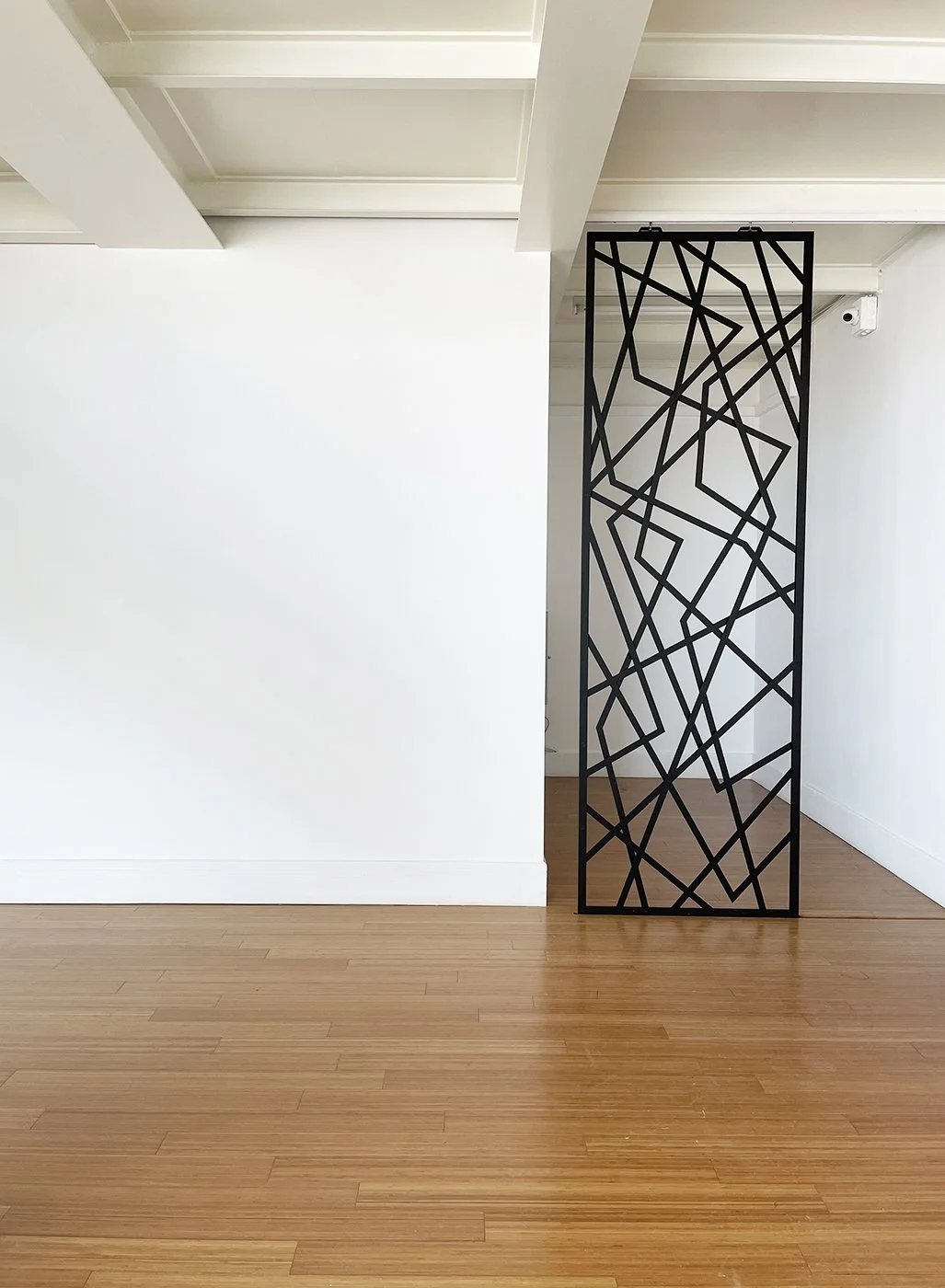 A black metal decorative room divider with an abstract geometric pattern, positioned in a corner of a minimalistic room with white walls and hardwood floors.
