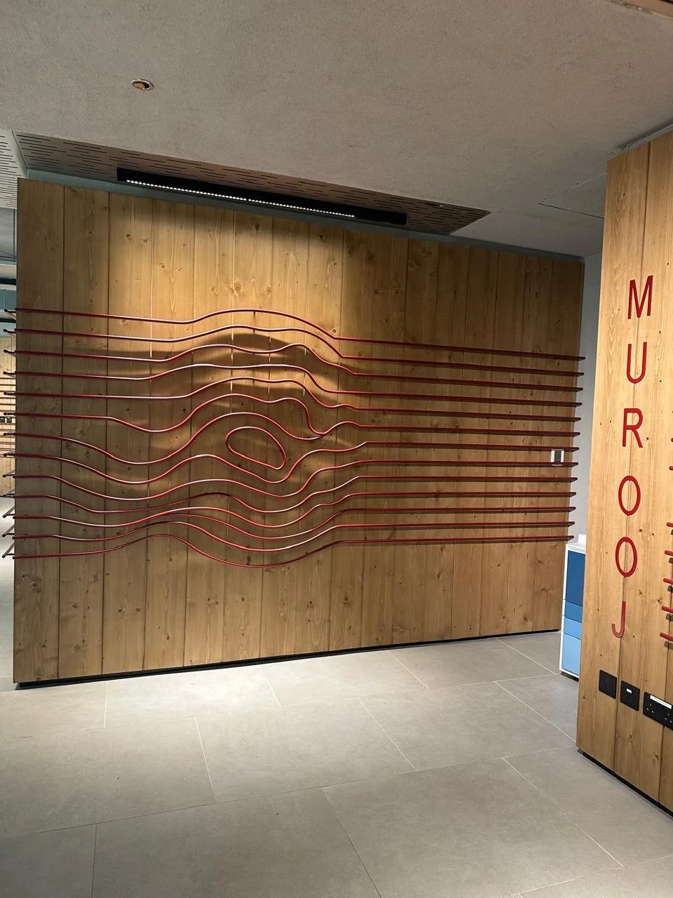 Interior wall with horizontal red metal rods creating a wave pattern on a wooden panel, and the word 'MURROJ' vertically in red on a wooden column.