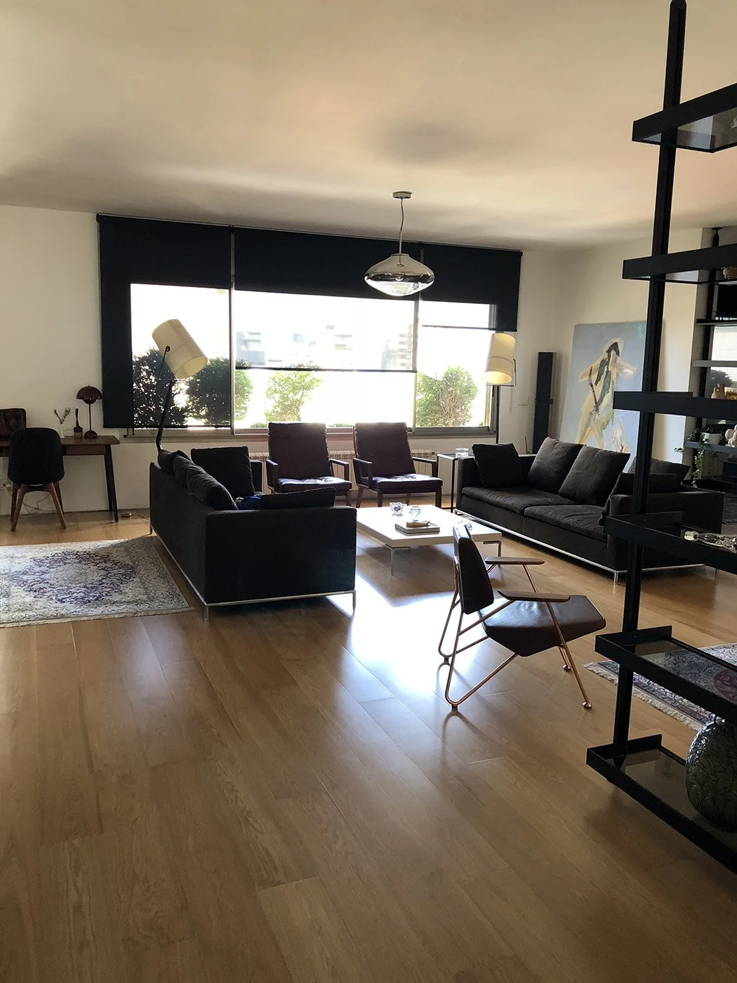 Bright living room with large window, black sofas, wooden chairs, white coffee table, desk with chair, bookshelf, and modern art painting.