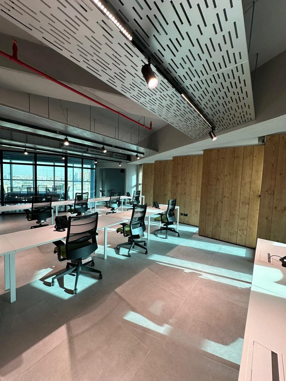 Empty modern office conference room with multiple white desks, black ergonomic chairs, black telephones, large windows with city view, wood paneling on one wall, and a perforated acoustic ceiling with track lighting.