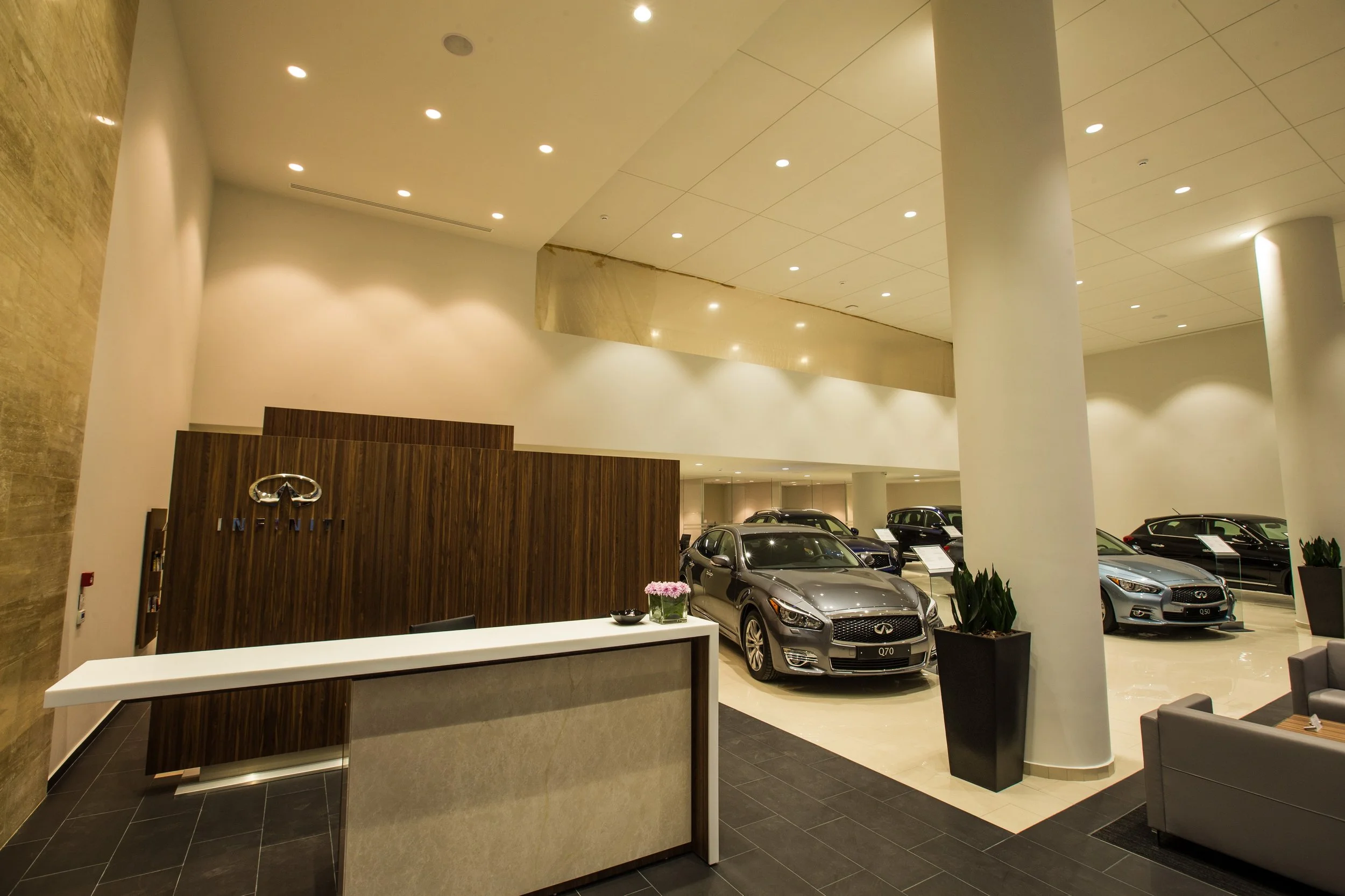 Interior of a car dealership with multiple luxury cars on display, tall white pillars, a curved ceiling with recessed lighting, a reception desk, and decorative plants.