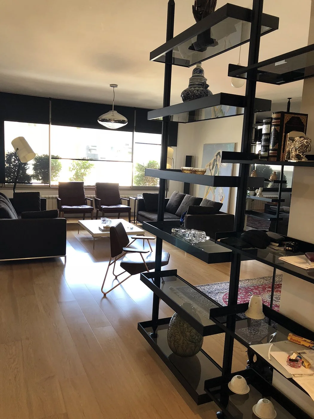 Living room with black sofas, wooden floor, large windows with dark blinds, and various decorative items on a black shelving unit.