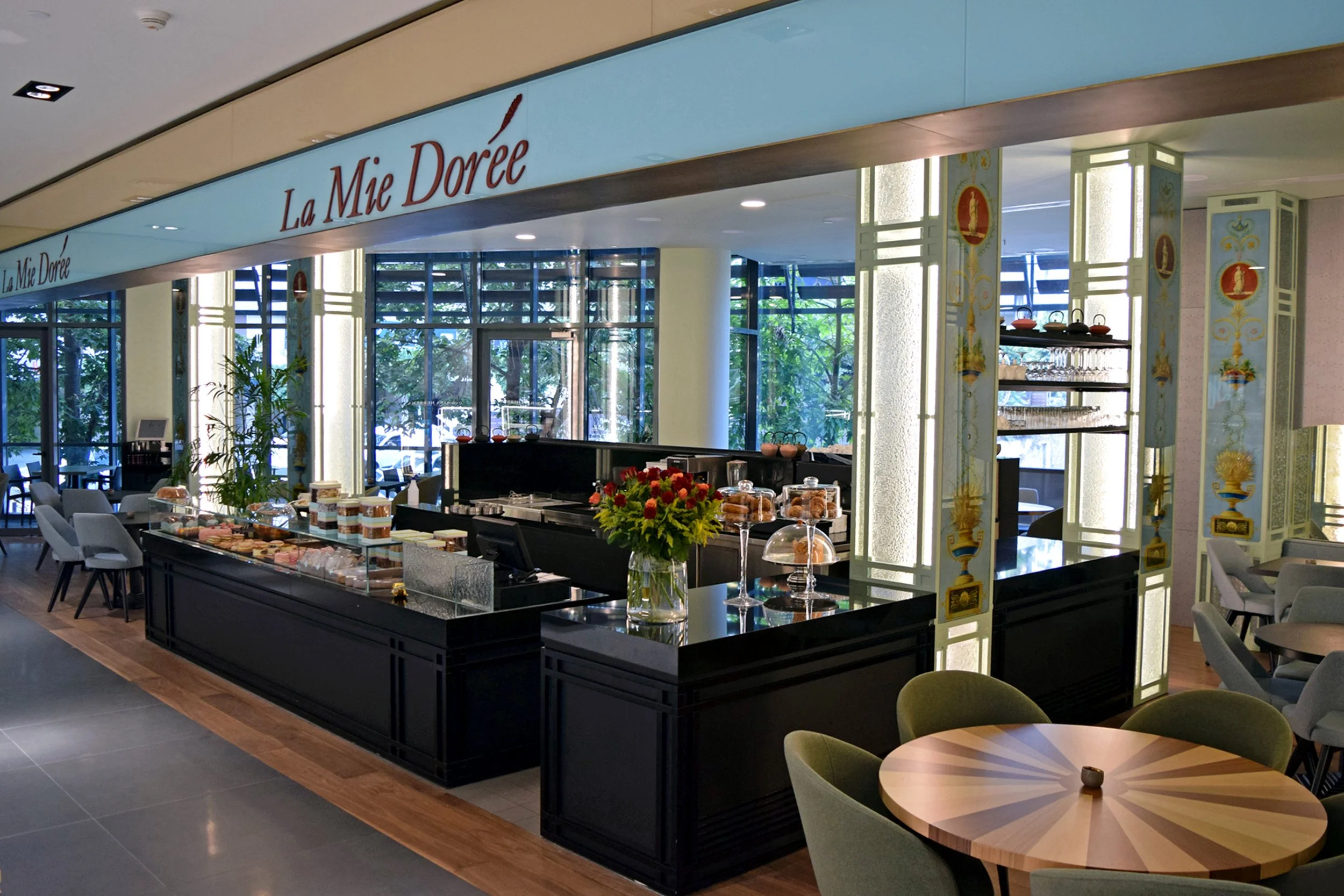 Interior of a bakery or café named La Mie Dorée with a pastry display counter, tables, and chairs, decorated with floral arrangements and illuminated by natural light from large windows.