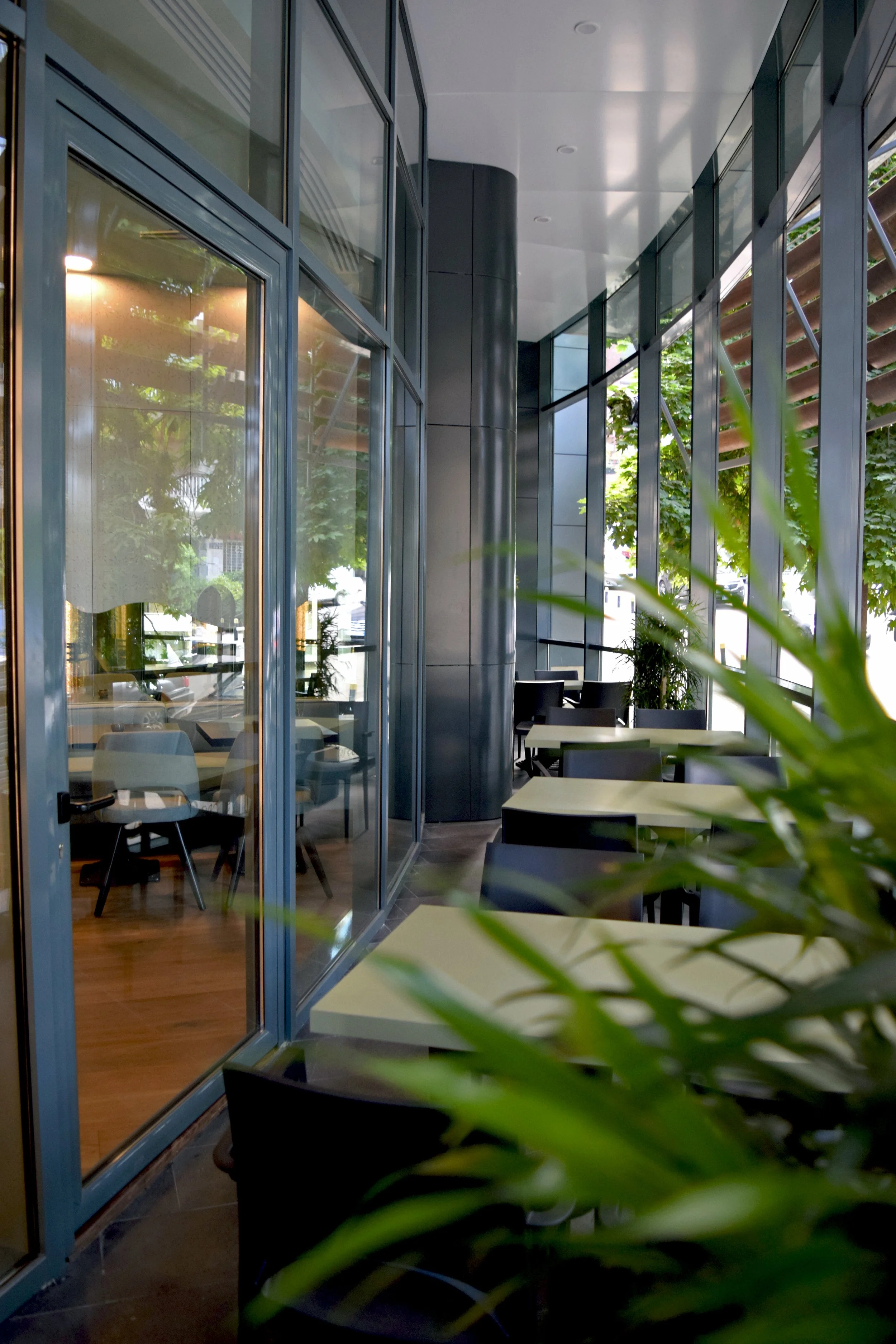 Modern indoor-outdoor workspace with glass walls, tables, chairs, and greenery outside.