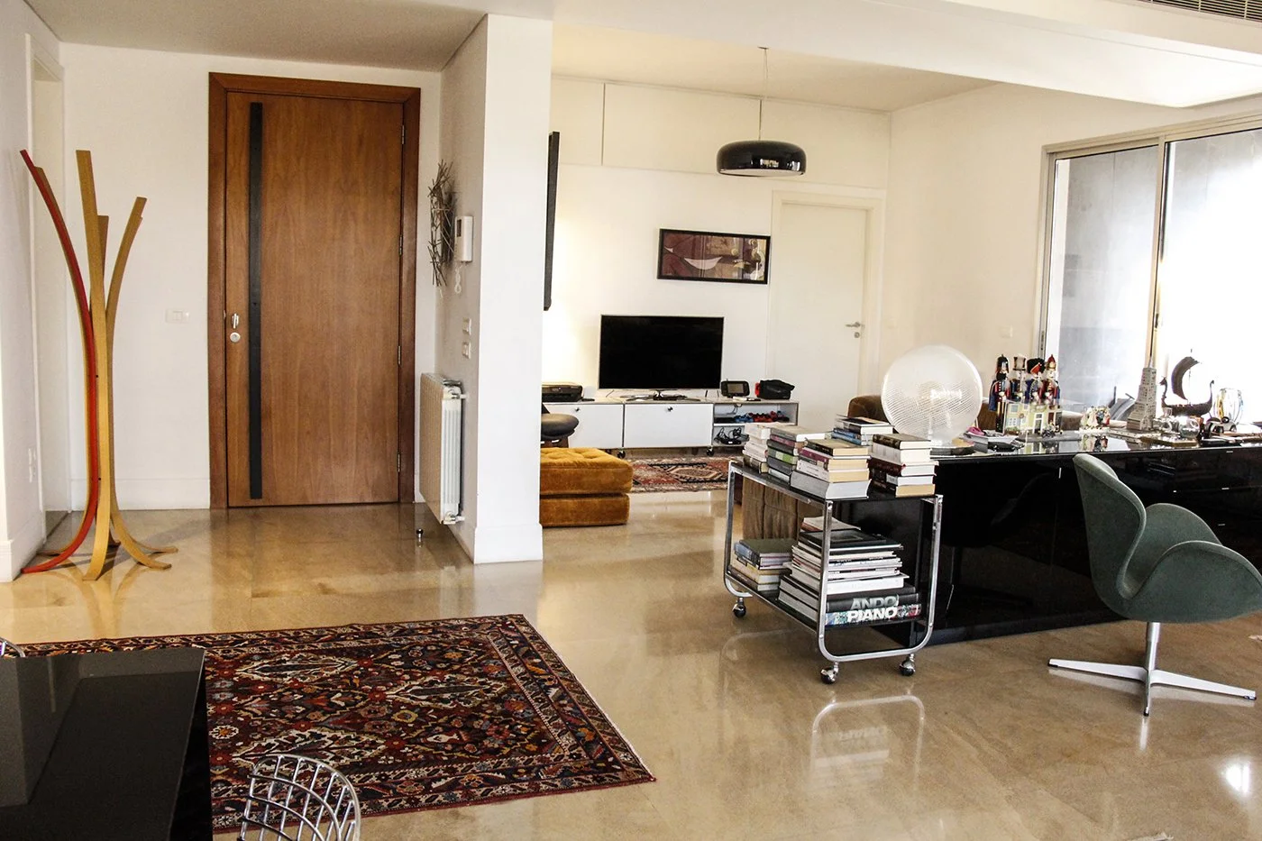 Living room with a wooden front door, a coat stand, a patterned rug, a television, a stacked book cart, a green swivel chair, a glass table with books, and large windows.