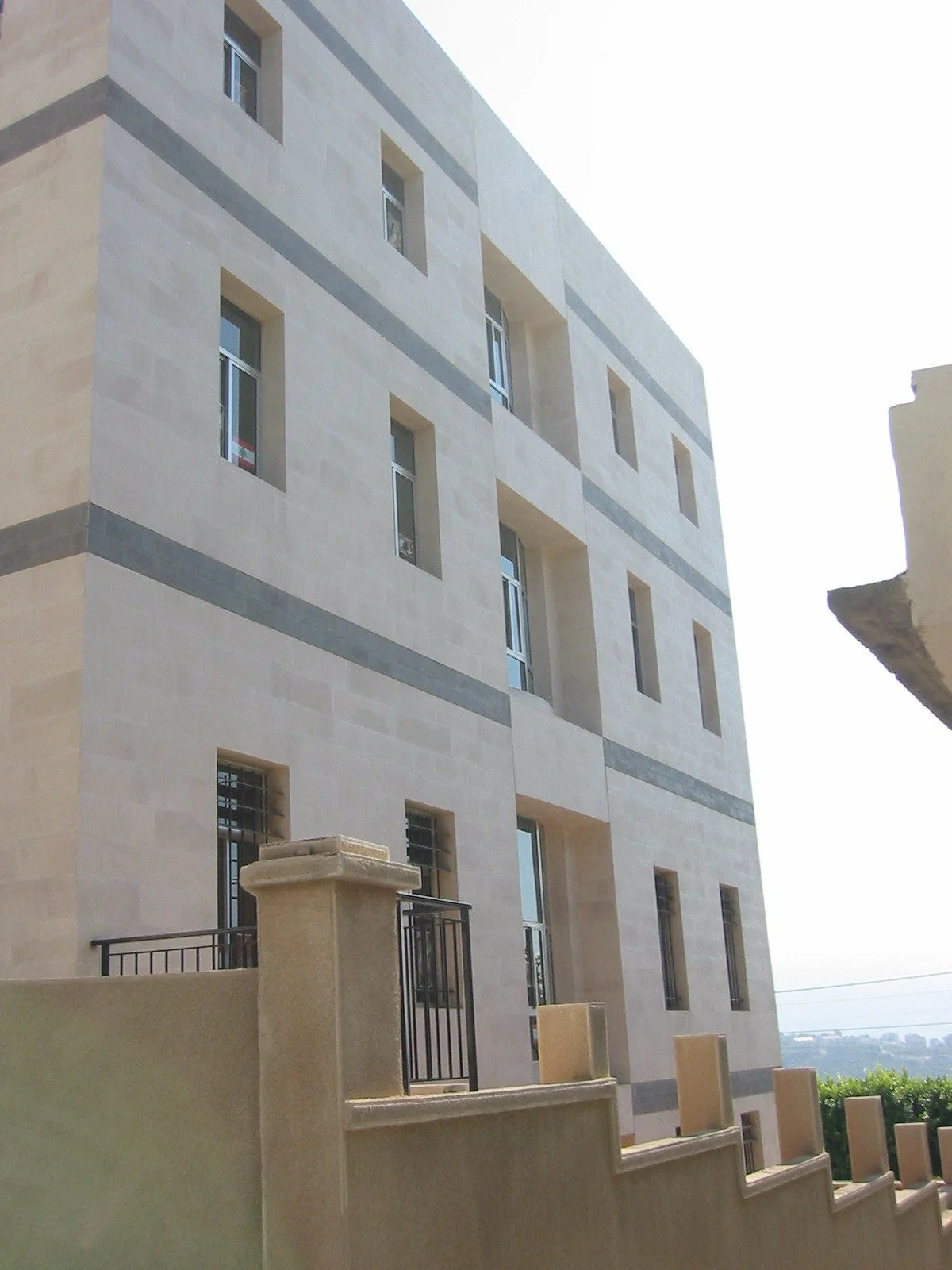 A modern multi-story beige building with rectangular windows and gray accents, featuring a small balcony with a black railing in the foreground.