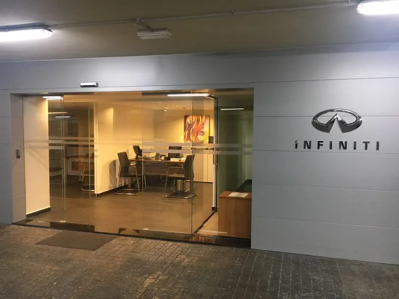 Exterior of an Infiniti dealership with a glass entrance and showroom visible inside. The Infiniti logo and name are on the wall to the right of the entrance.