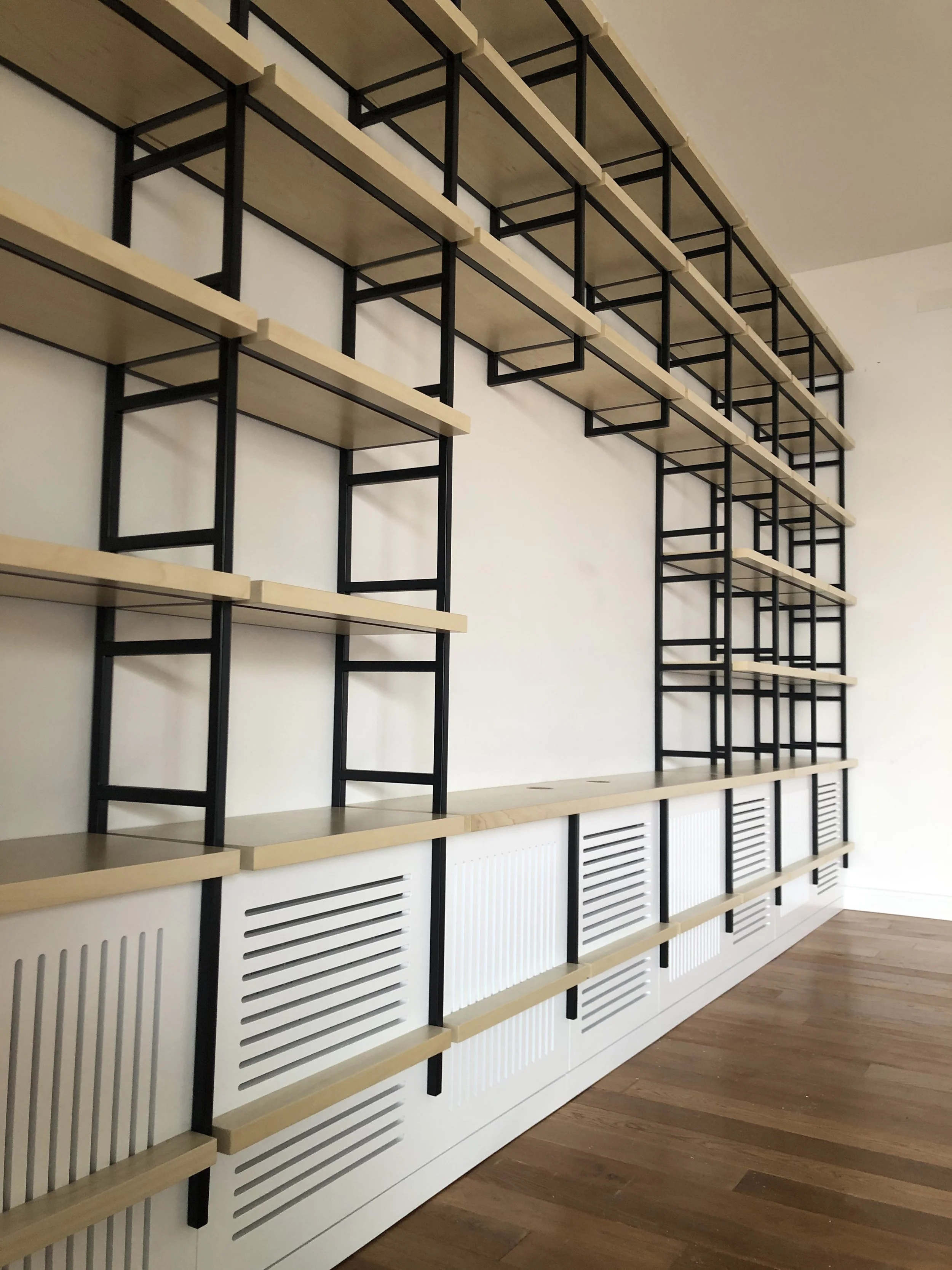 Wall-mounted shelving unit with wooden shelves and black metal supports, located in a room with white walls and wooden flooring.