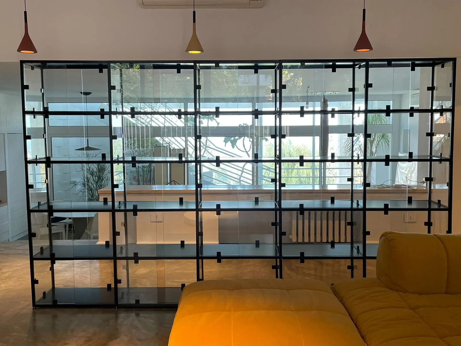 Interior view of a modern room with a black metal and glass shelving unit, yellow sofa, wooden floor, and large windows in the background with plants and trees outside.