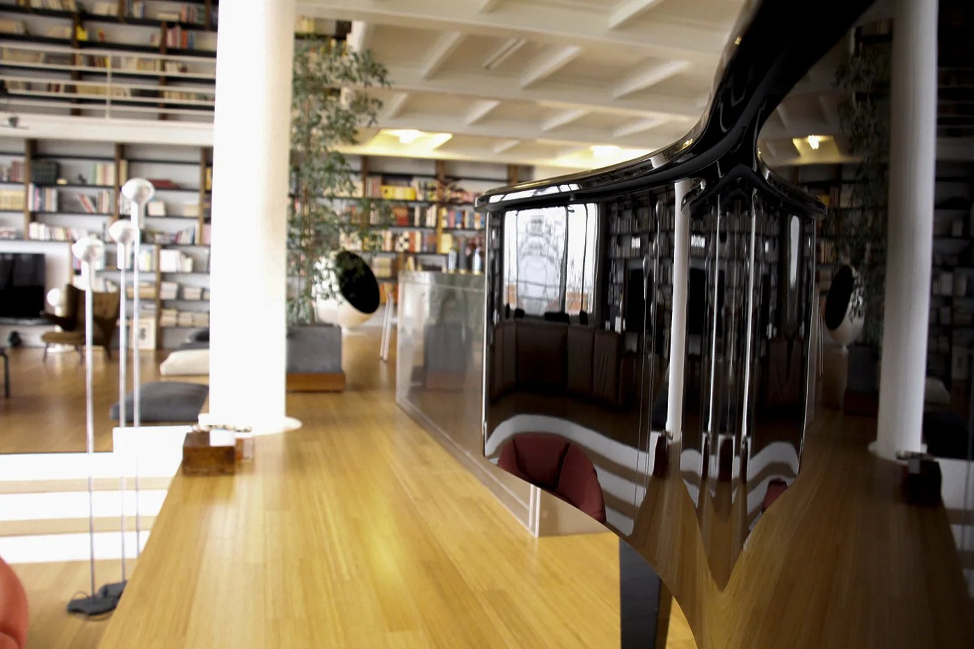Reflection of a house and furniture in the polished black surface of a grand piano, with a background of bookshelves in a modern, well-lit room.
