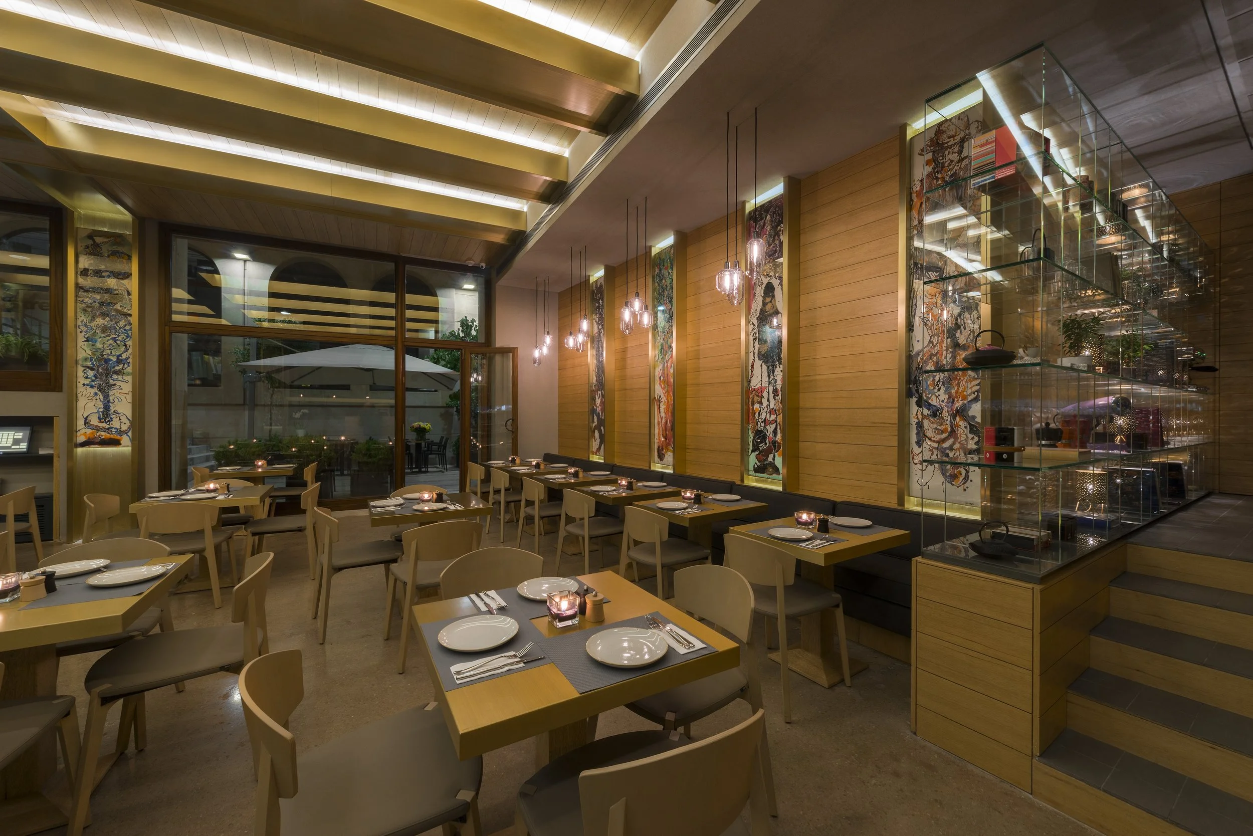 Interior of a modern restaurant with wooden walls, large glass windows, and hanging pendant lights. Dining tables are set with plates, napkins, and candles. Artistic paintings decorate the walls, and there is a glass display shelf with decorative ite
