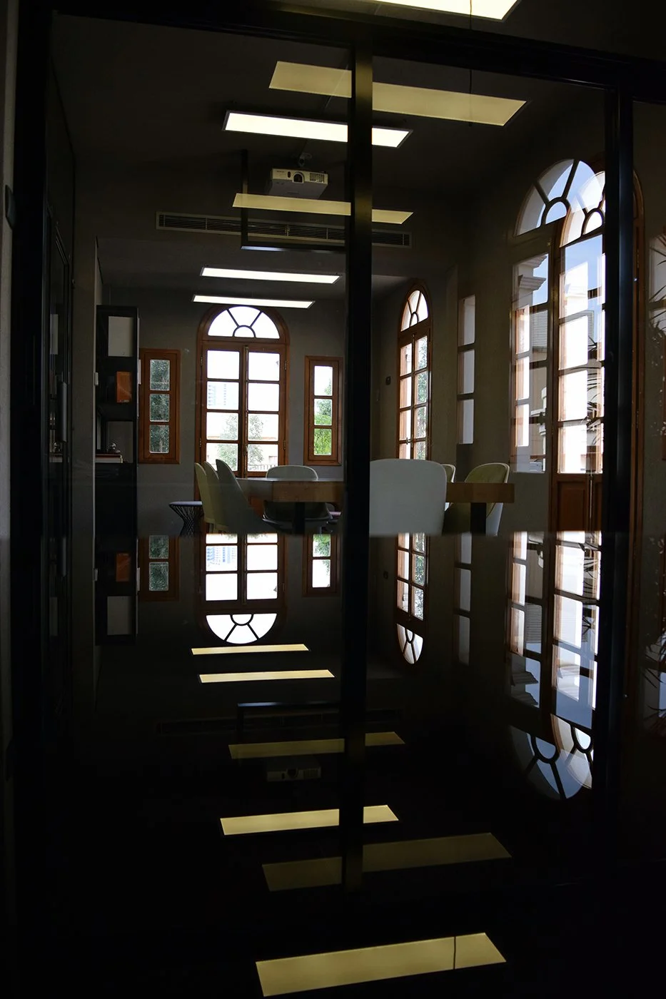 View of a conference room with large arched windows, a wooden table, and chairs, seen through glass doors with reflections of the room's interior.