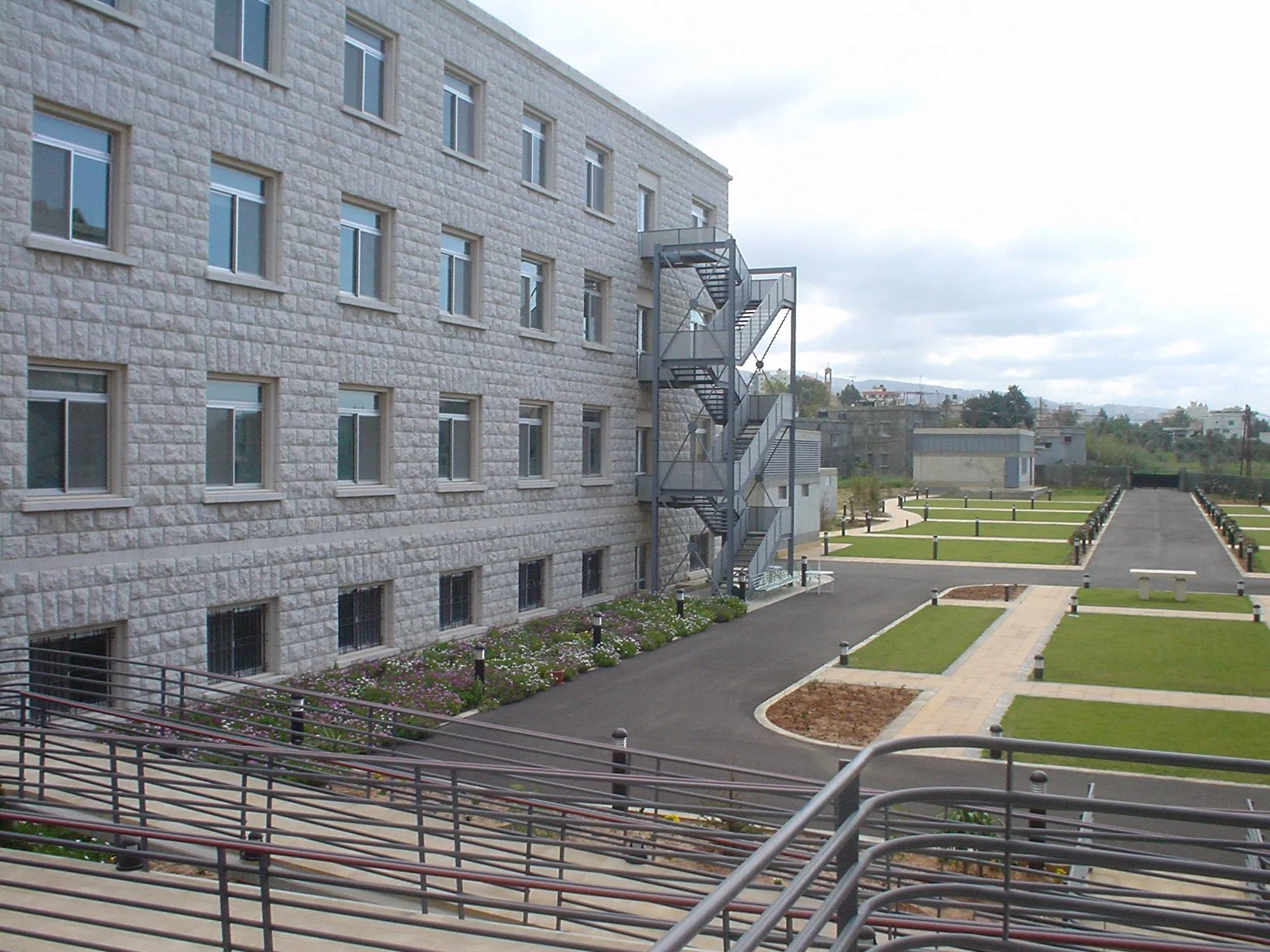 Exterior view of a multi-story building with a stone facade, fire escape stairs on the side, and a landscaped courtyard with pathways, flower beds, and benches.