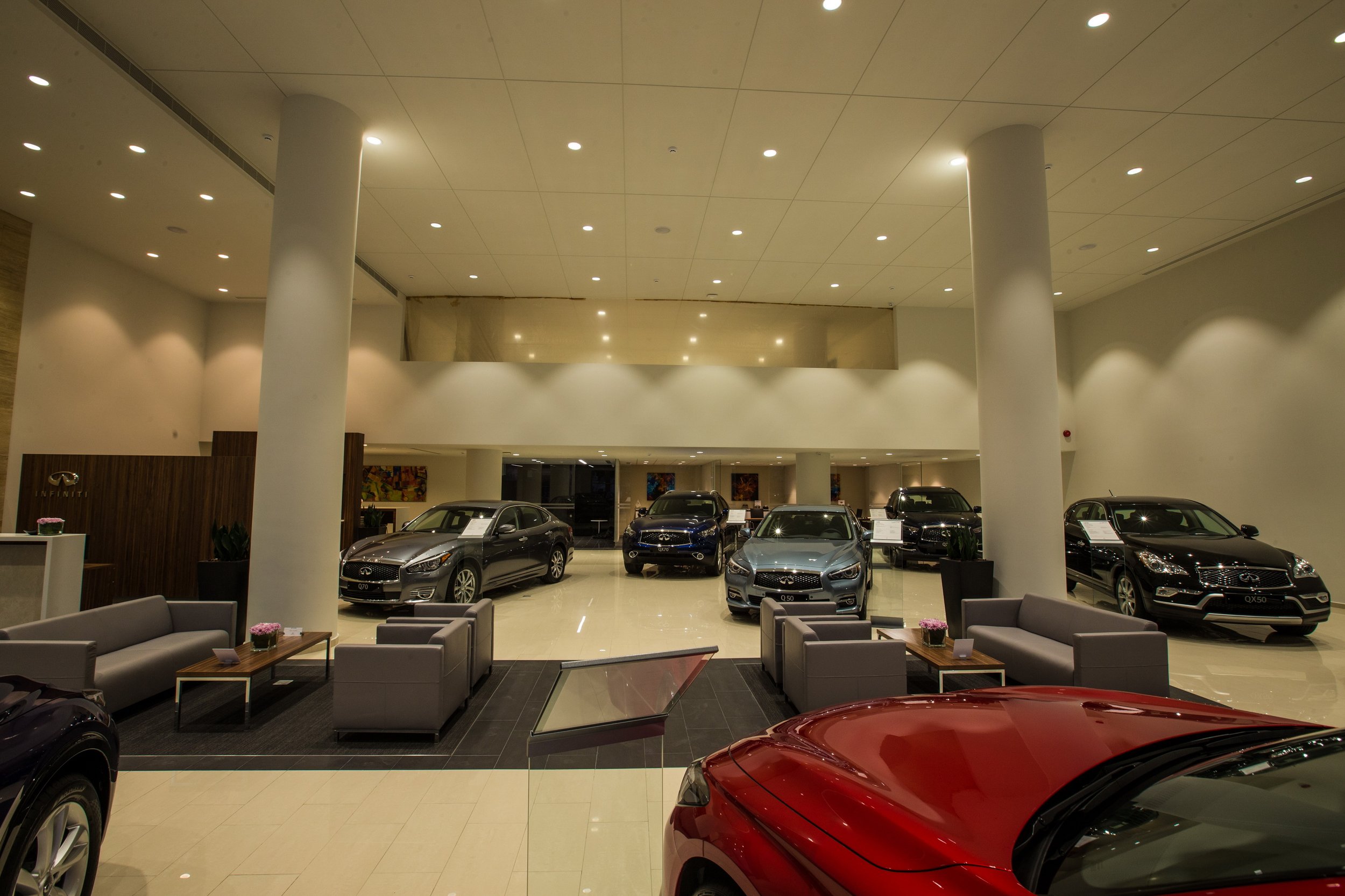 The image shows the interior of a car dealership with several new Infiniti cars on display, a seating area with sofas and tables in the foreground, and a large open space with a high ceiling and white columns in the background.
