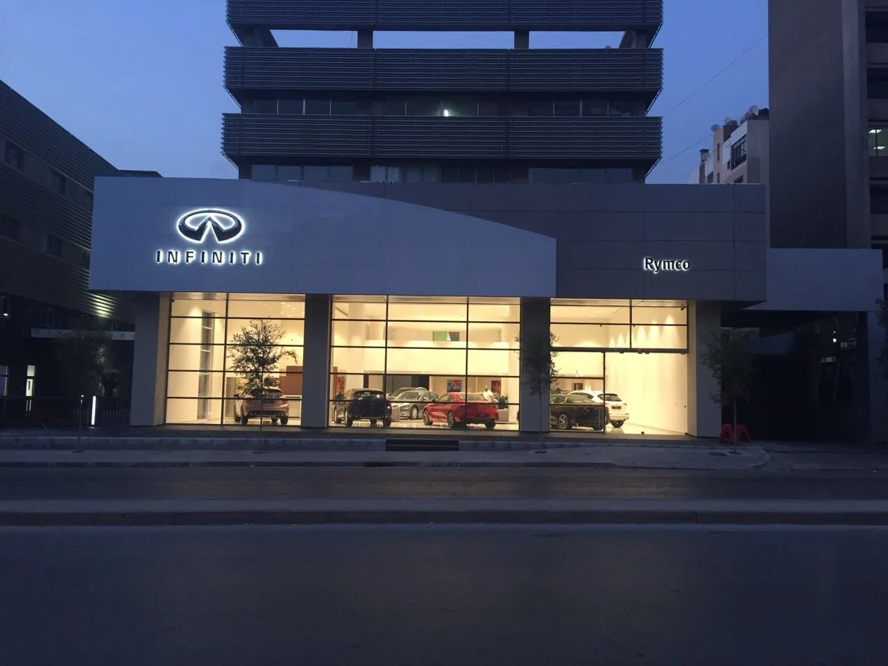 Exterior view of an Infiniti car dealership during evening with illuminated signage and cars inside the showroom.