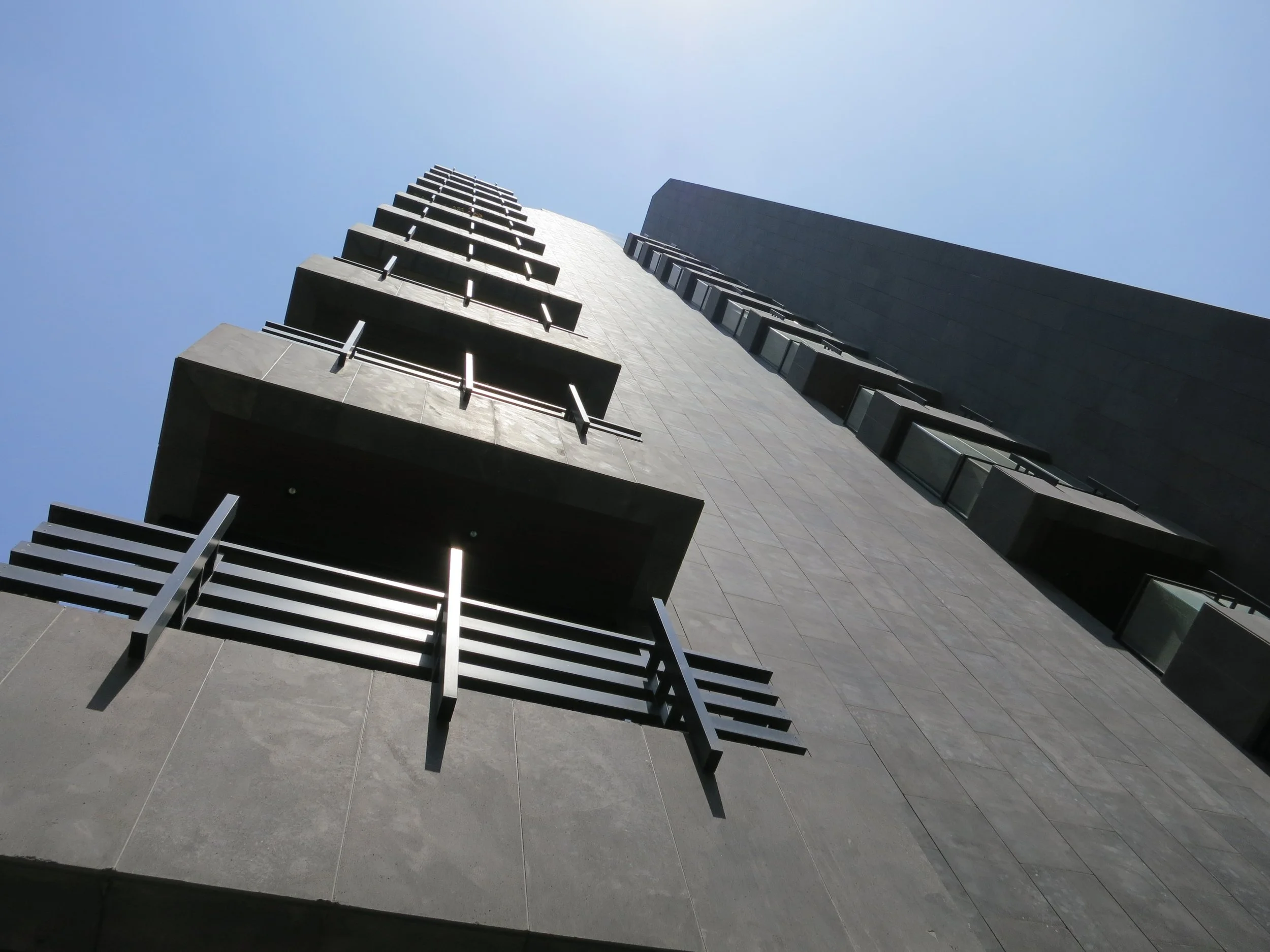 A tall modern building seen from the ground looking upward, with balconies and windows, against a clear, blue sky.