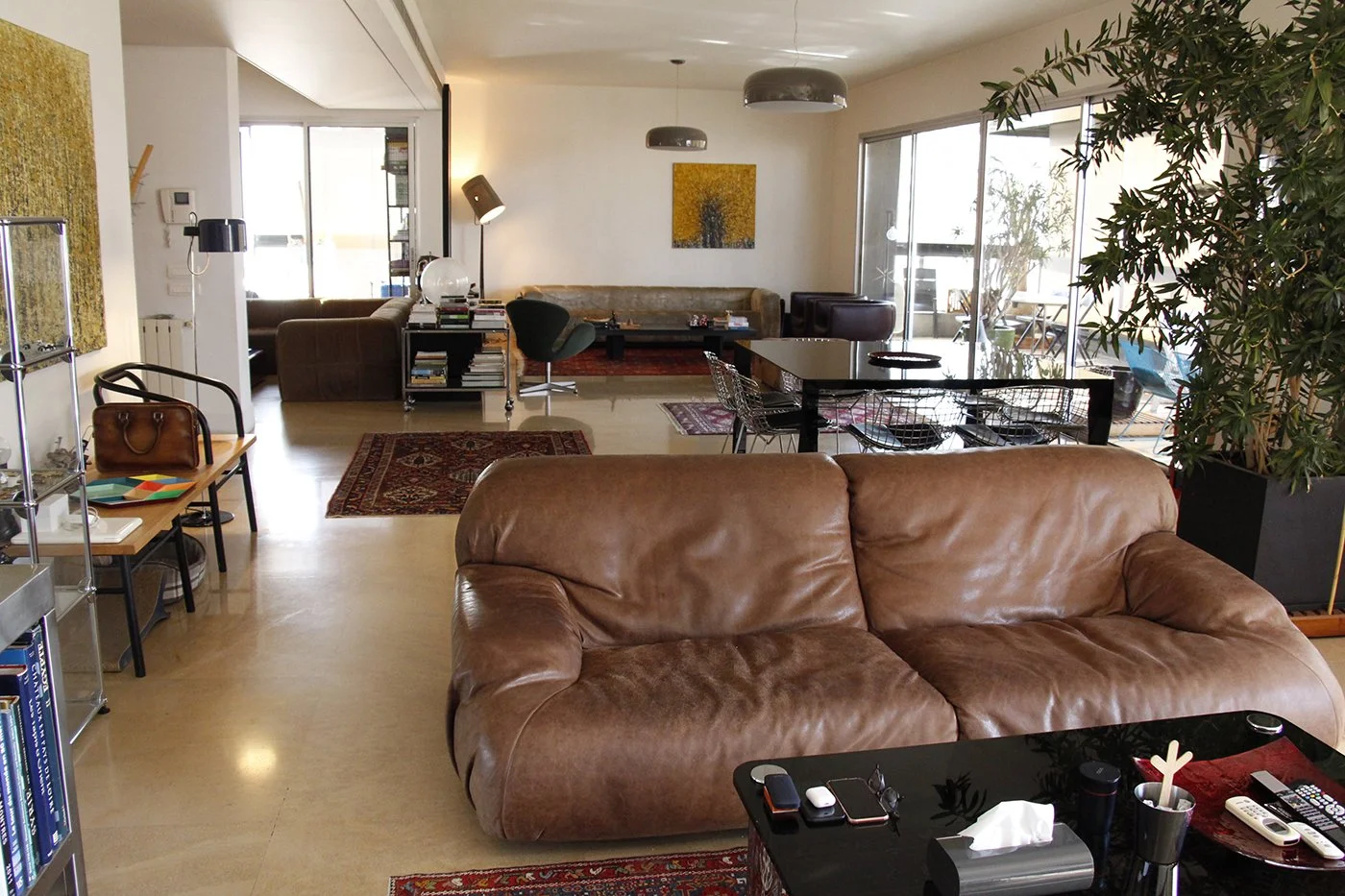 A spacious living room with a brown leather sofa, various chairs, rugs, potted plants, and a dining table with chairs. Glass doors lead to a balcony with outdoor furniture.