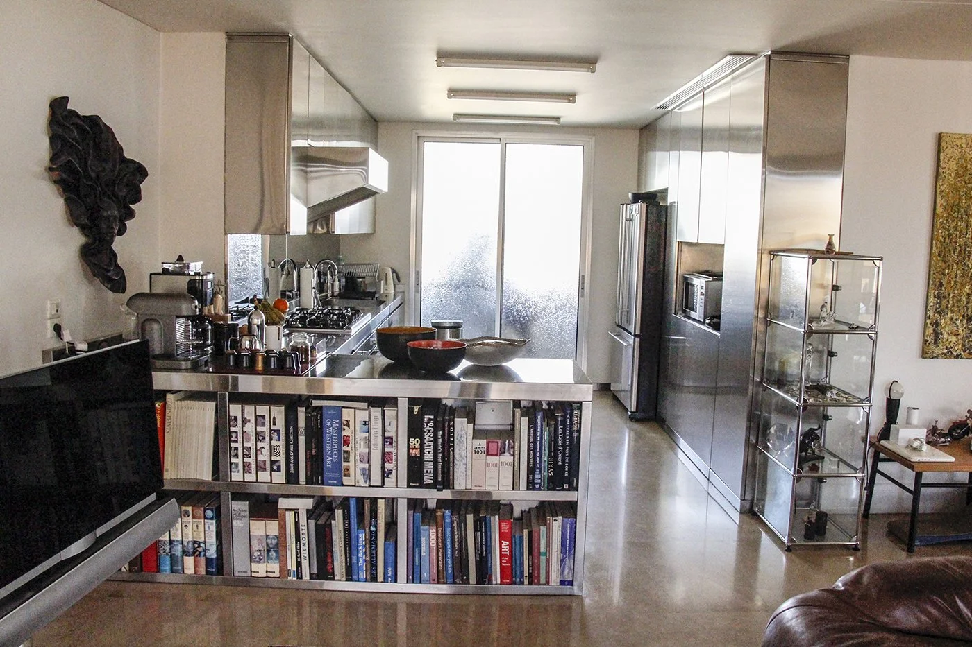 Modern kitchen with stainless steel appliances, a sliding glass door, a bookshelf with cookbooks, and a metal display cabinet filled with collectibles.