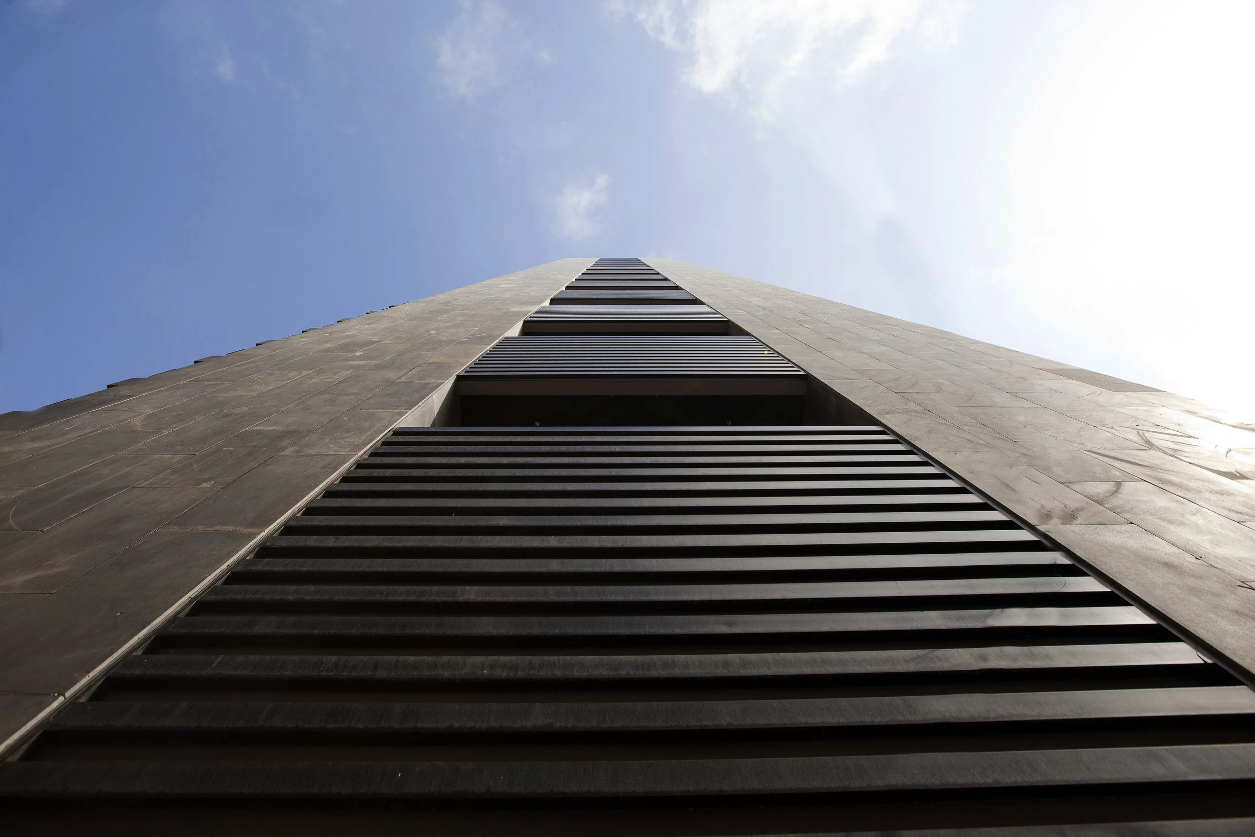Low-angle view of a tall modern skyscraper with black horizontal louvers and a clear blue sky in the background.
