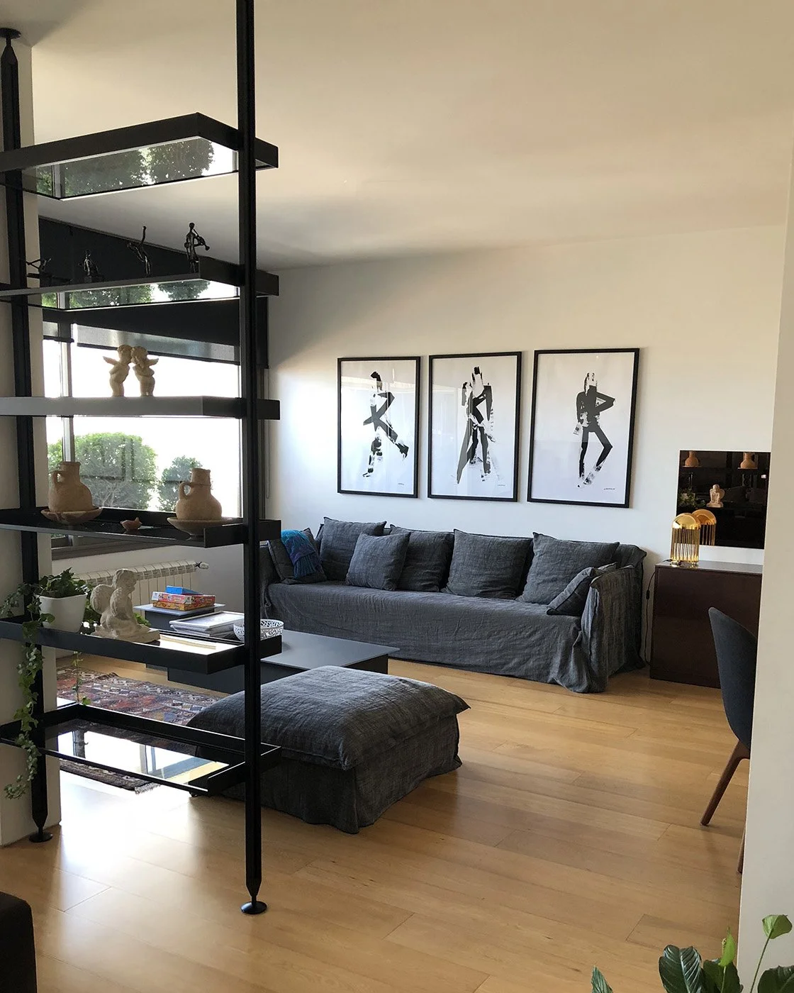 Living room with gray sofa, black and white artwork on wall, wooden floor, and black metal open-shelving unit with decorative items.