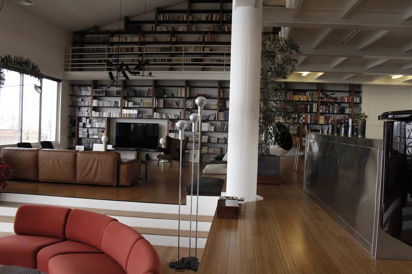 A spacious living room with large windows, a brown leather sofa, a red sectional sofa, and a wall of bookshelves filled with books. There is a chandelier, a flat-screen TV, and a tall plant. The room features hardwood floors and modern furniture.