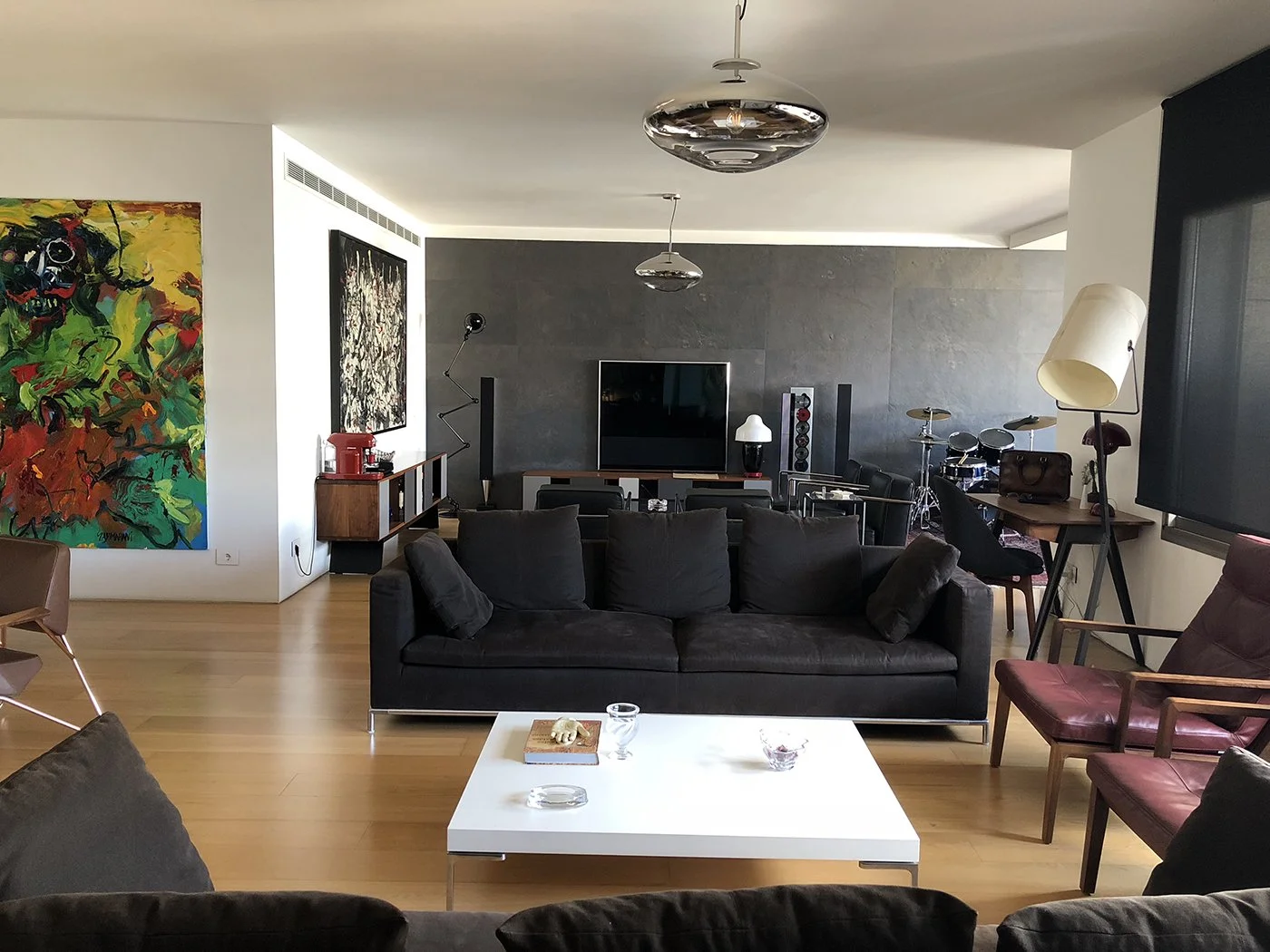 Living room with black sofa, white coffee table, colorful abstract paintings, drum set, television, desk with lamp and bag, armchair, floor lamp, and artwork.