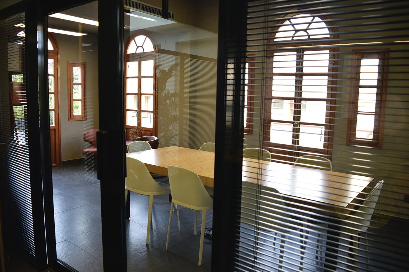 An interior conference room seen through glass panels with blinds, featuring a wooden table, white chairs, and large arched windows letting in natural light.