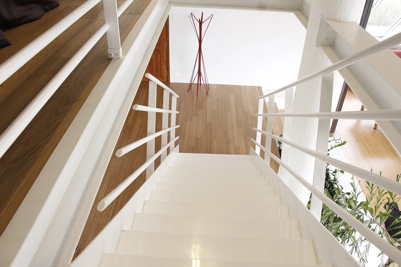 Top view of a staircase with white steps and white railings leading to a wooden floor, with a red coat rack and a doorway visible in the background.