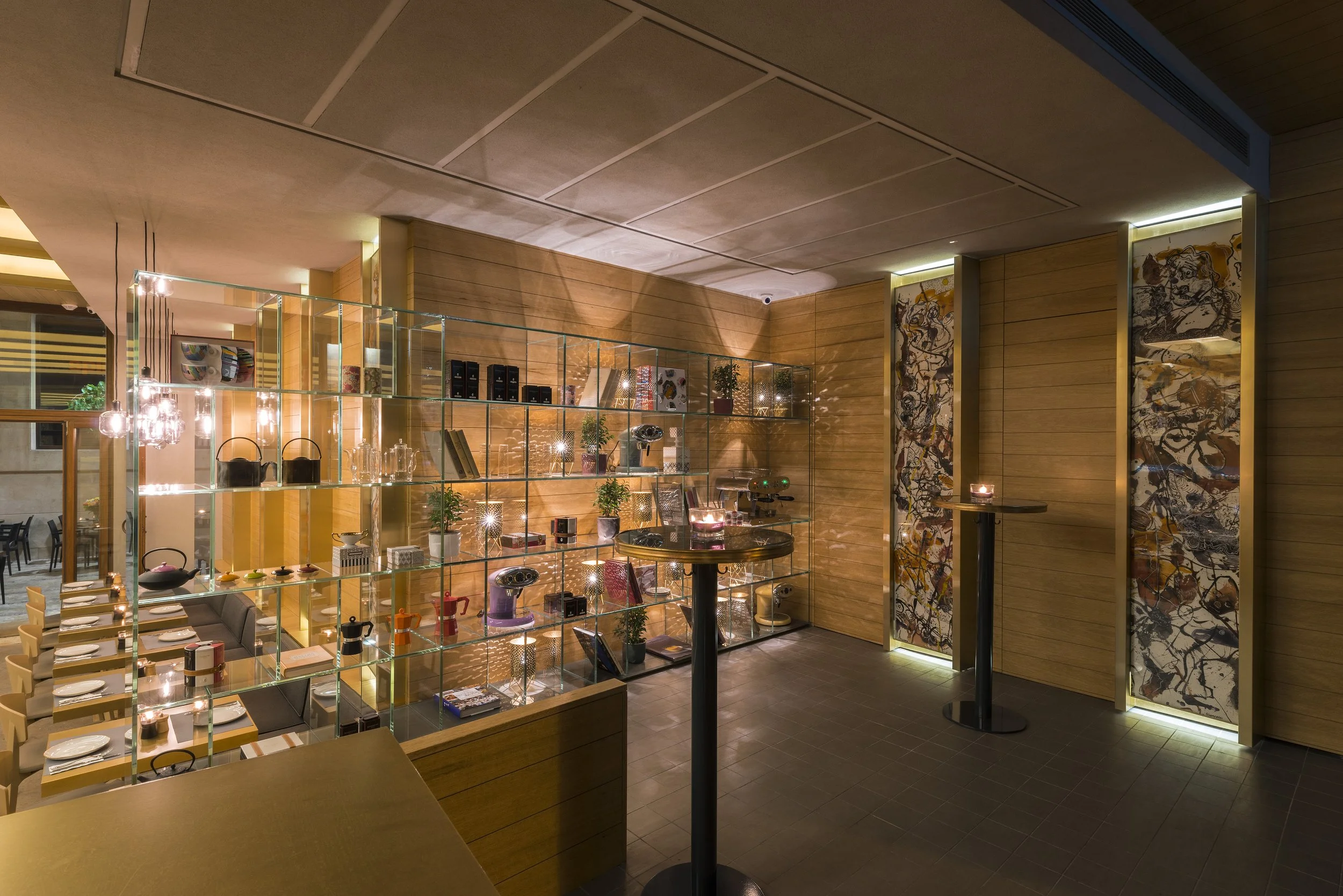 Interior of a modern restaurant or café with wooden walls, art pieces, and a glass shelving unit displaying kitchenware and decorative items. The space is softly lit with warm lighting.