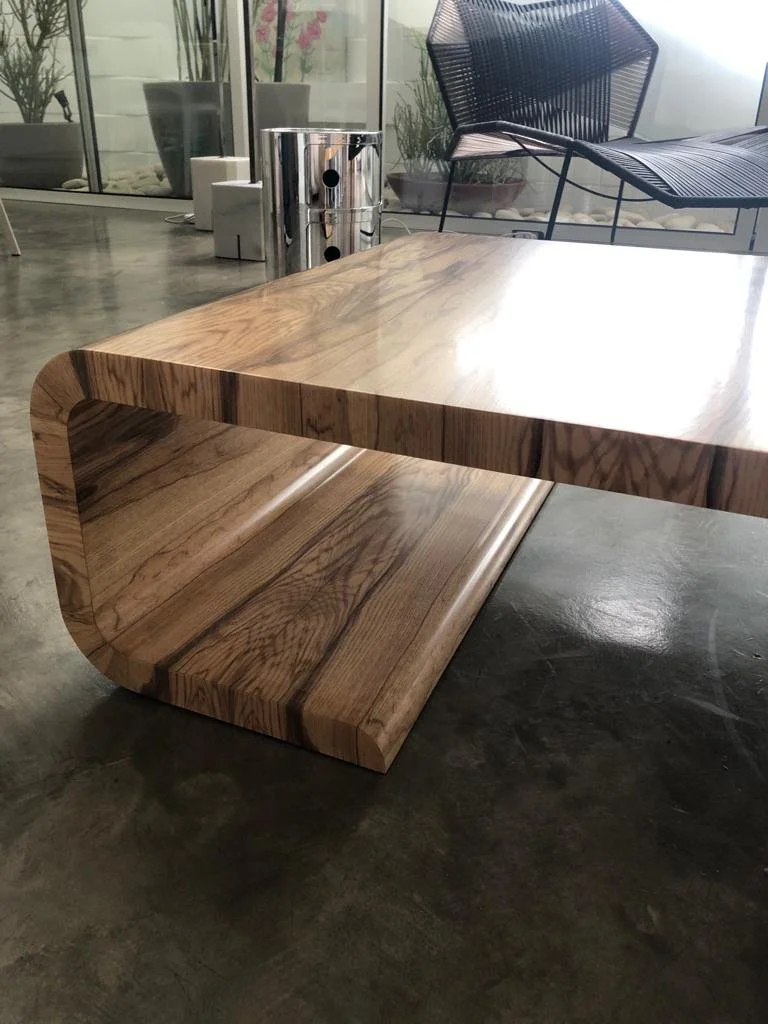 Modern wooden coffee table with curved edges in a contemporary living space with glass walls, potted plants, and black wire chair.