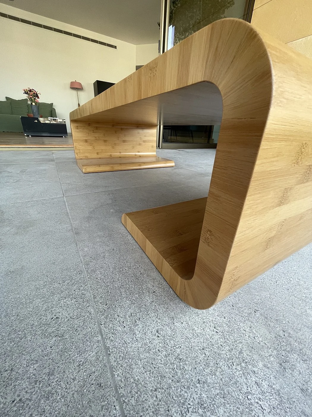 Modern wooden coffee table with a curved design, placed on a tiled floor in a living room with a green sofa and a pink lamp in the background.