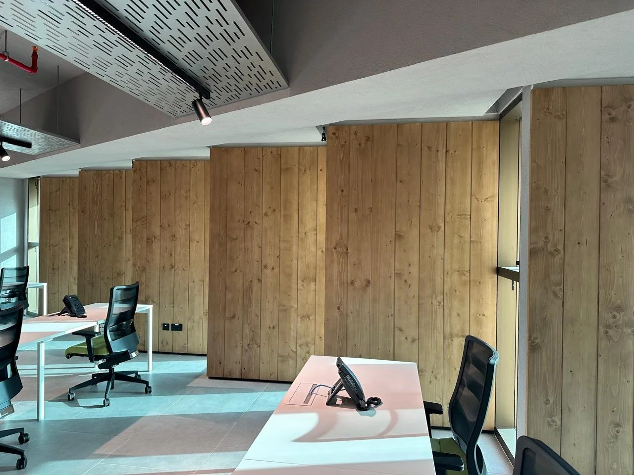 Empty modern office with white desks, black office chairs, and wooden wall panels, with windows and ceiling lights.