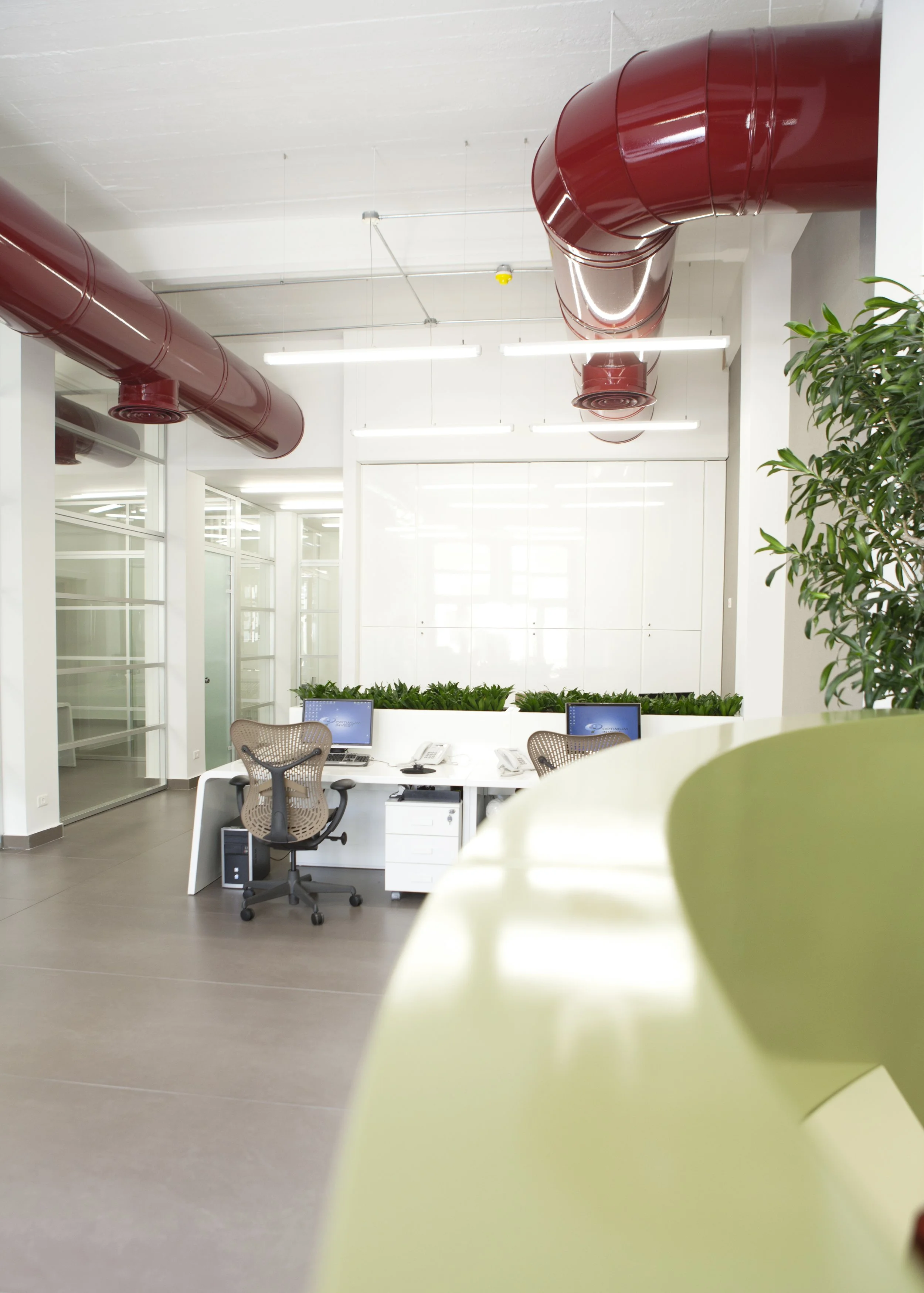 Modern office space with two desks, each with a computer, phone, and office chair. There are green plants behind the desks, bright lighting, and large red ventilation ducts on the ceiling. A yellow object is partially visible in the foreground.