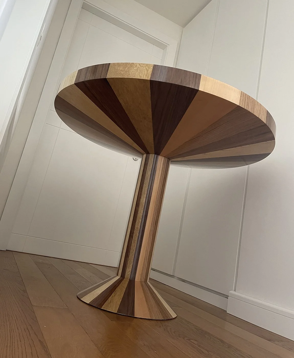 A modern wooden side table with a round top and a pedestal base, made of wood panels in varying shades of brown.