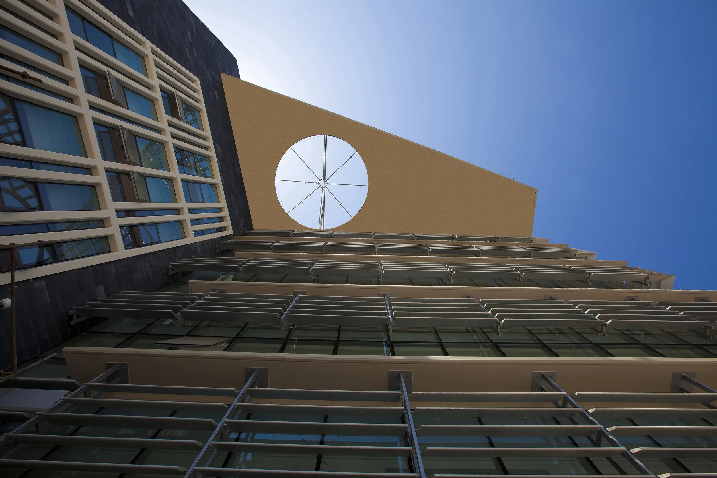 Looking up at a modern multi-story building with glass windows and balcony railings, featuring a large circular structure in the upper part of the building, with a clear blue sky in the background.