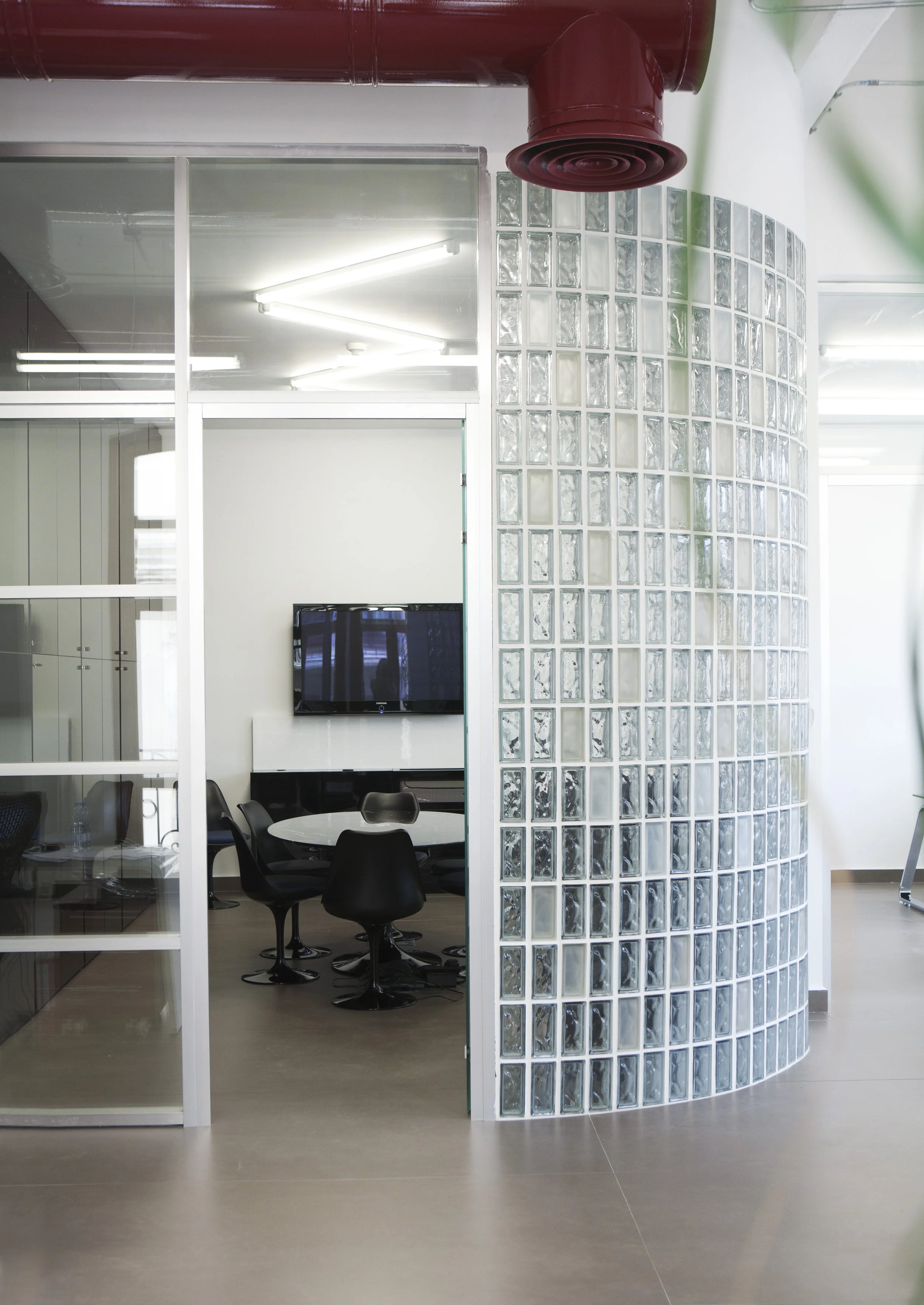 Modern office conference room with a round table and black chairs, glass wall with sliding doors, a large flat-screen TV, and a curved glass block wall with a red ceiling vent.