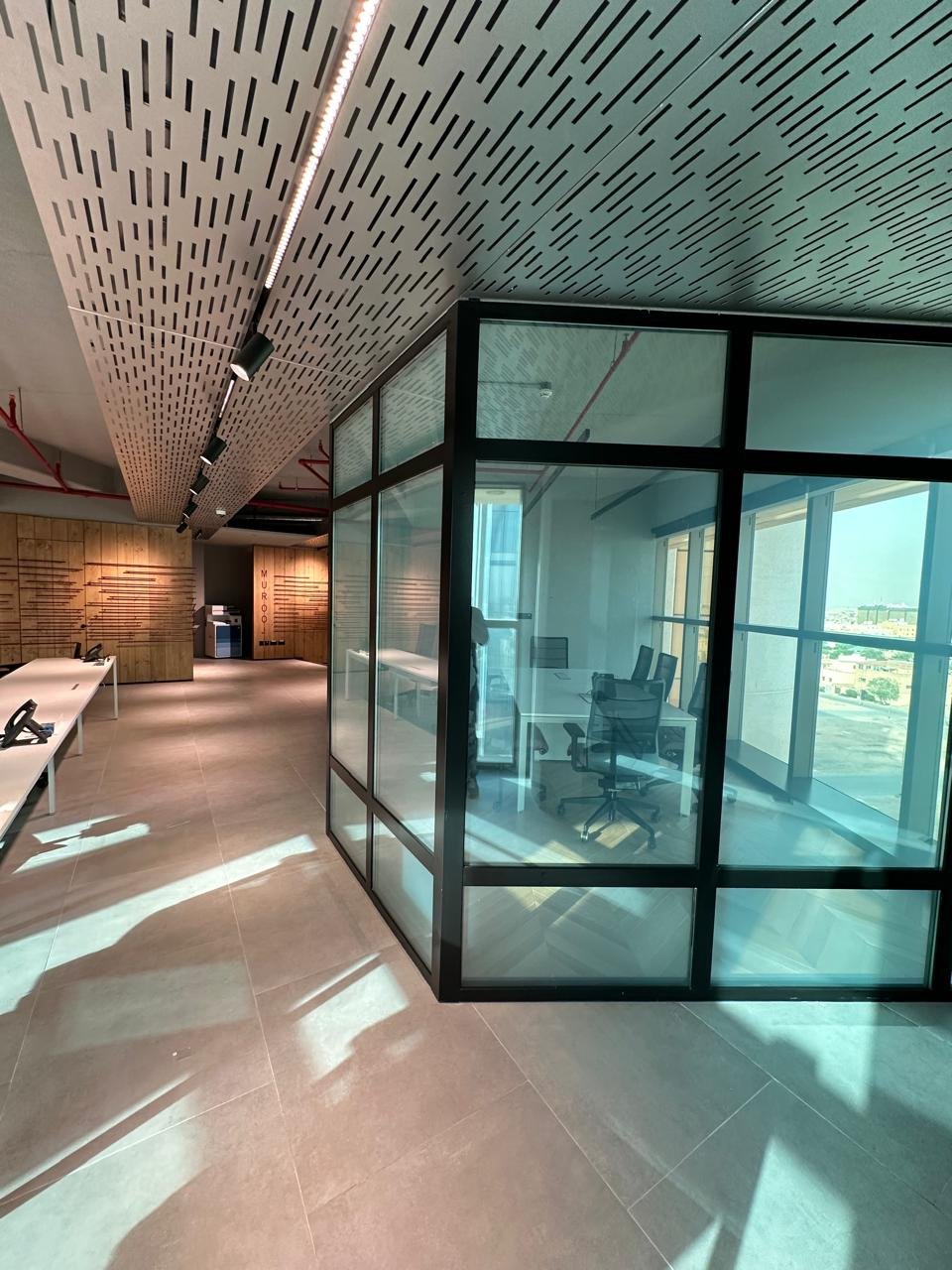 Modern office space with glass-walled conference room, wooden wall accents, and seating area with white benches, illuminated by natural sunlight.