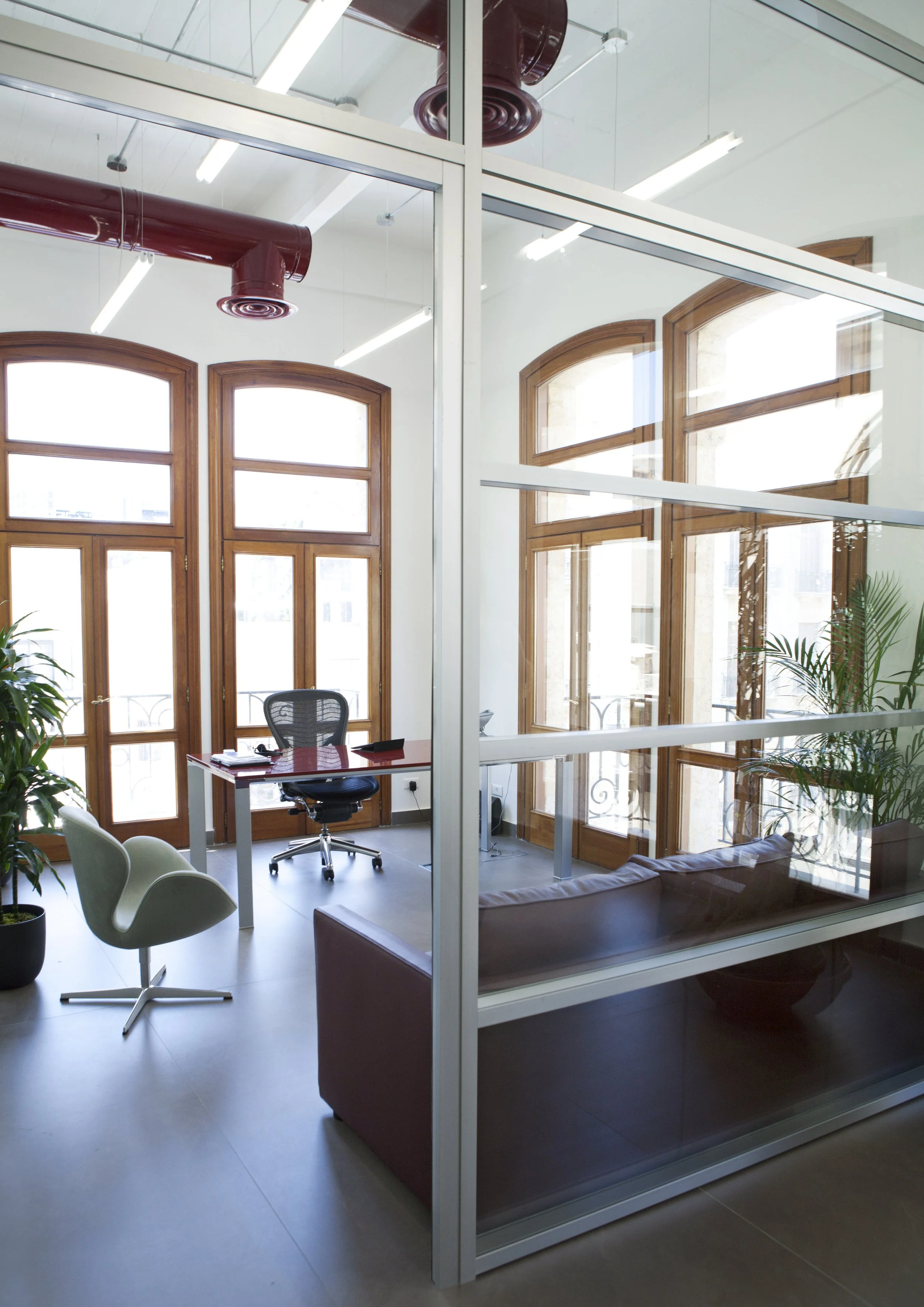 Modern office with large wooden framed windows, a desk with an ergonomic chair, a couch, and potted plants, separated by glass walls.