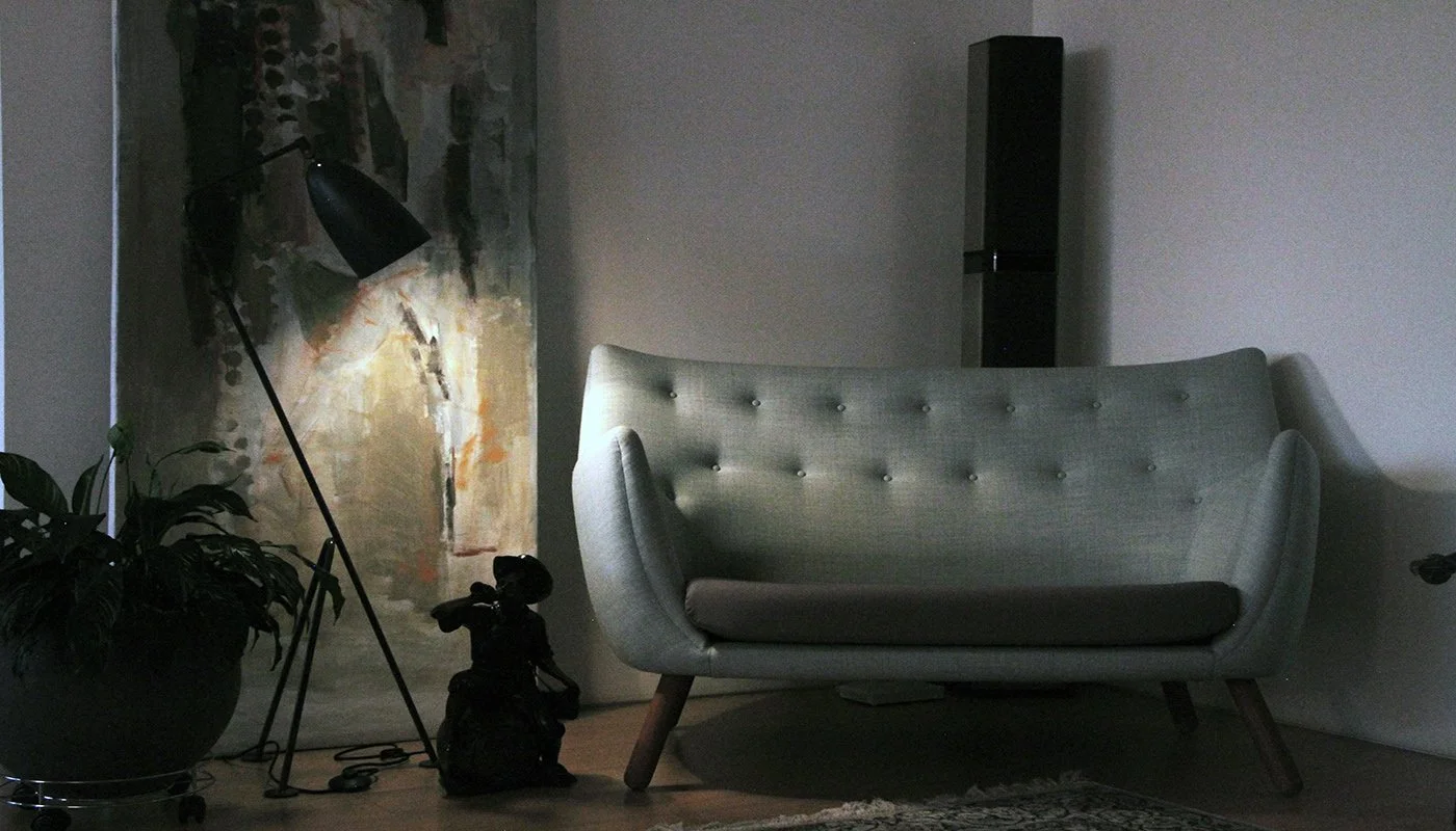 Living room corner with potted plant, floor lamp, abstract painting, gray sofa with wooden legs, and a small black speaker in the background.